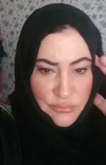 A woman with dark hair, wearing a black headscarf, posing with her hand near her face in front of a patterned background.