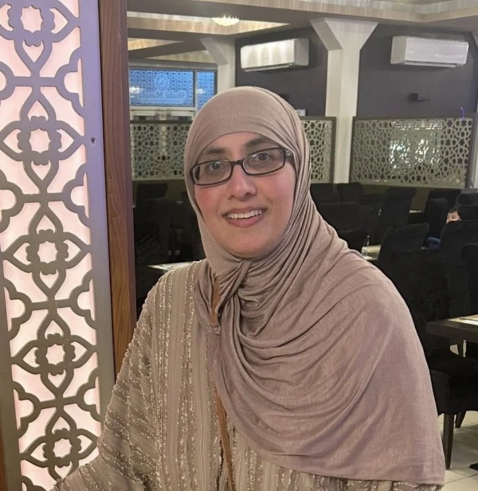 A woman wearing a beige hijab and glasses smiling in a restaurant with decorative wall patterns and dark chairs in the background.