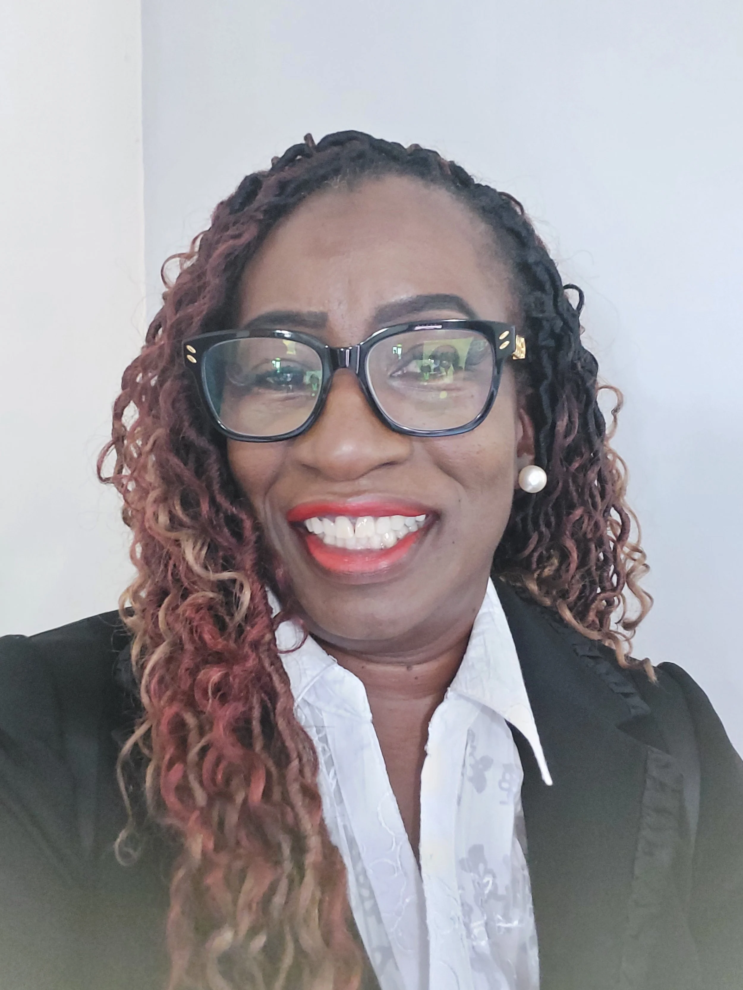 A smiling woman with curly hair, wearing glasses, pearl earrings, a white blouse, and a black blazer, posing for a selfie against a plain background.