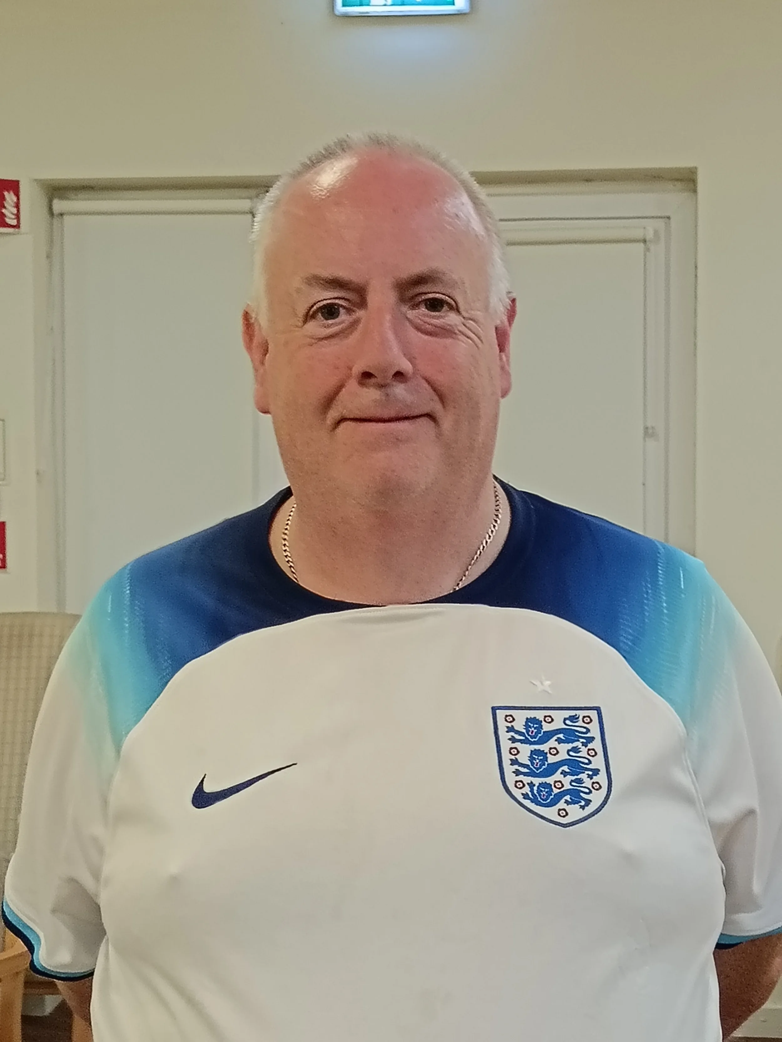 A man with short gray hair wearing an England football jersey with the national emblem and a Nike logo, standing indoors against a light-colored wall.