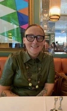 A woman wearing glasses and a green shirt, smiling at a restaurant table.