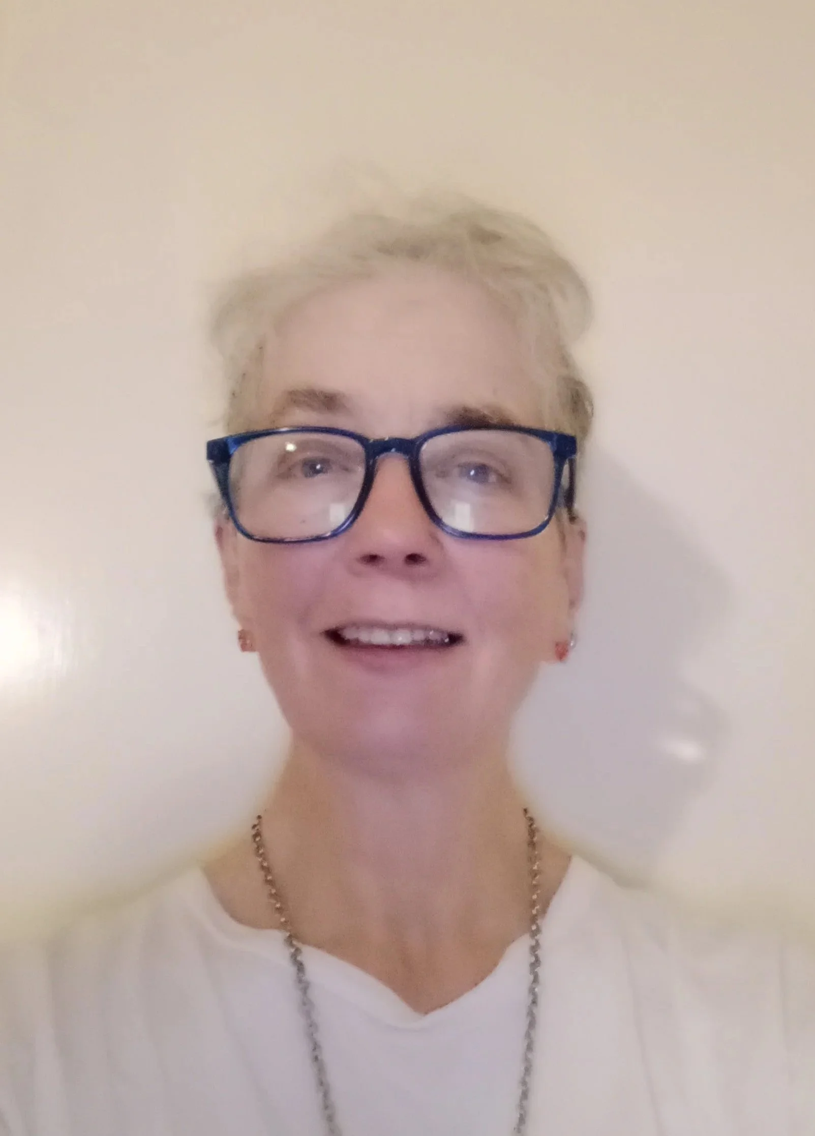 A woman with short gray hair, wearing glasses, earrings, a necklace, and a white shirt, smiling in front of a plain wall.