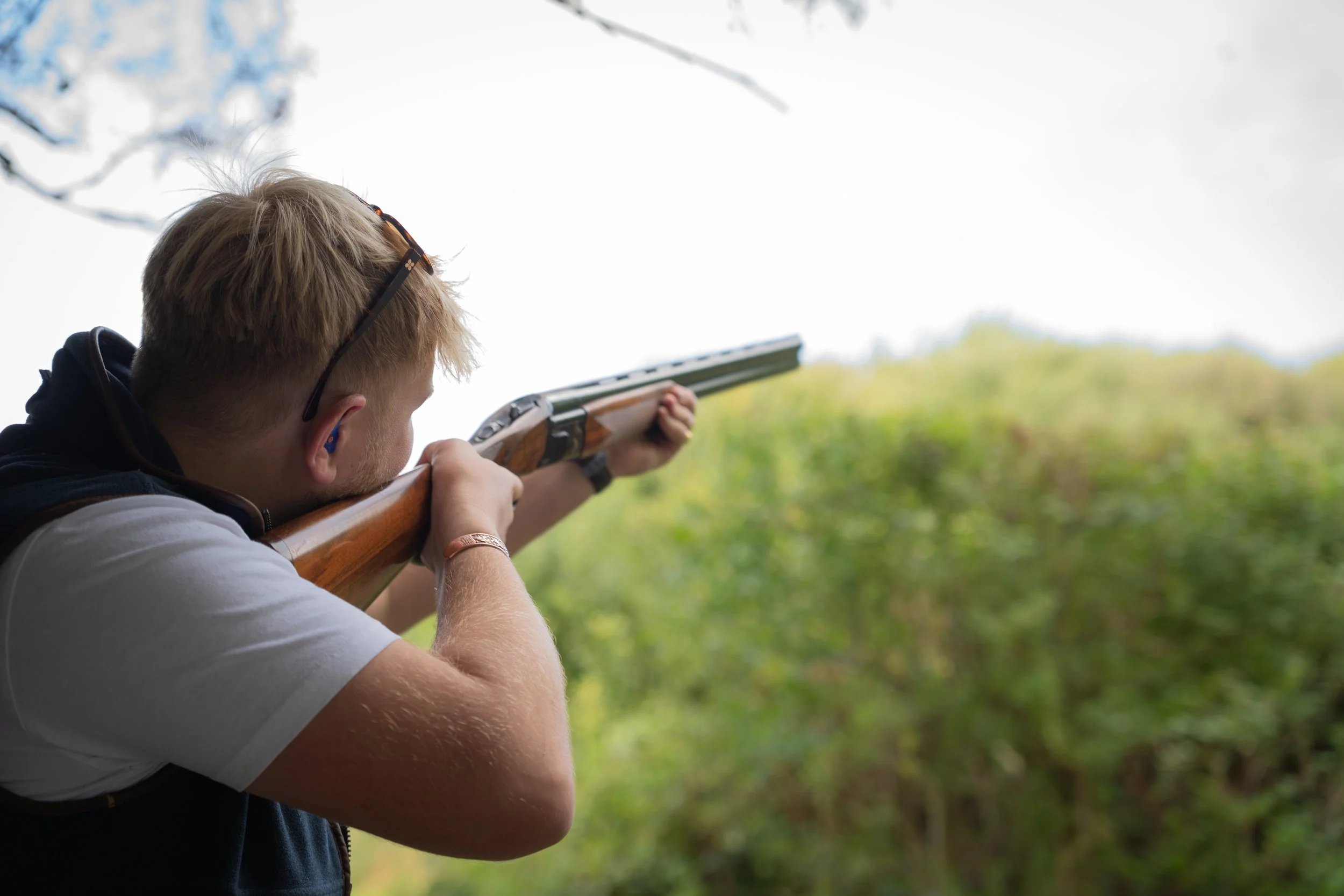A man aiming a shotgun outdoors with a forest in the background.