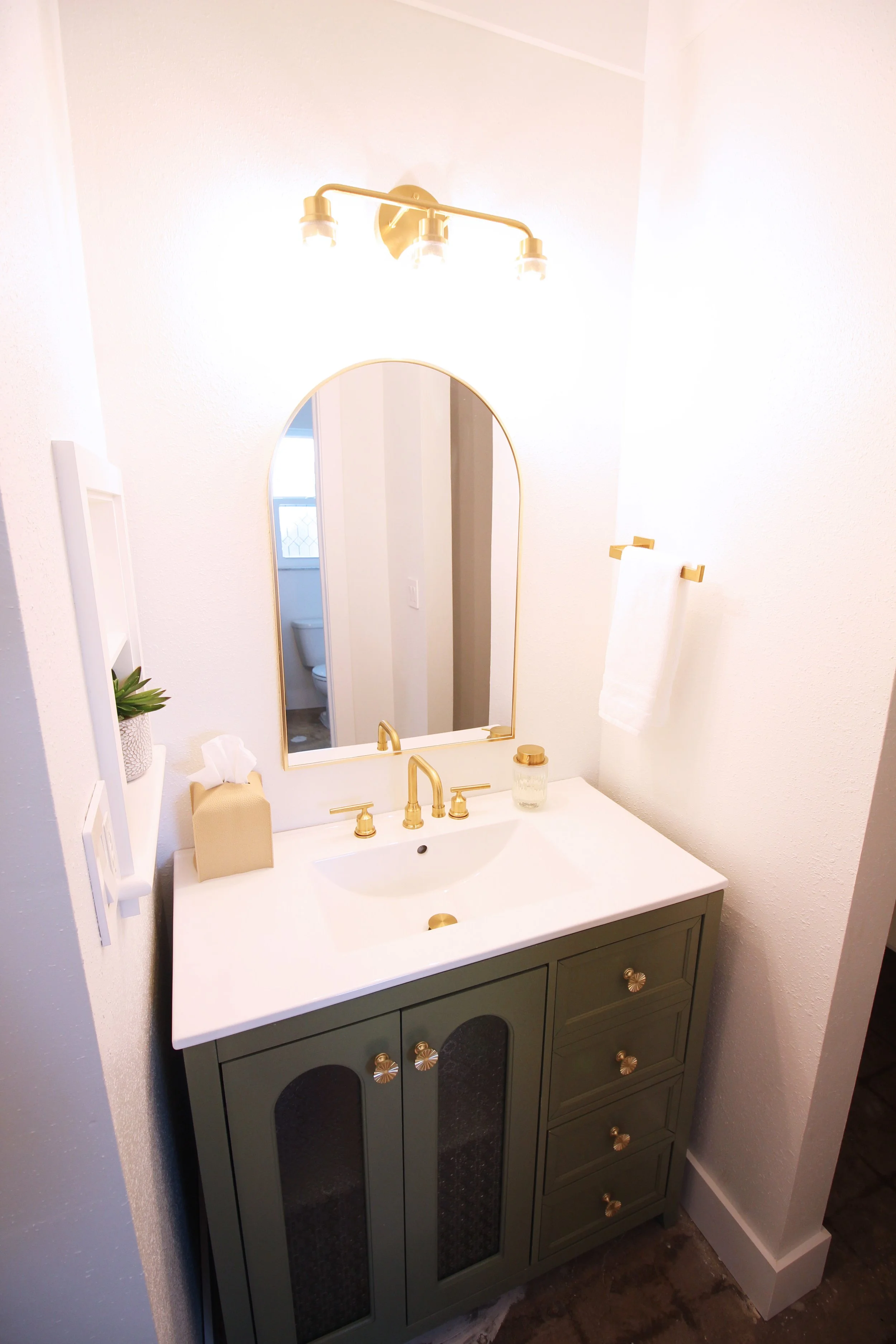 A bathroom with white walls, sage green vanity with a matte gold faucet, bright lighting, arched mirror, white countertop, tissue box, and a hand towel