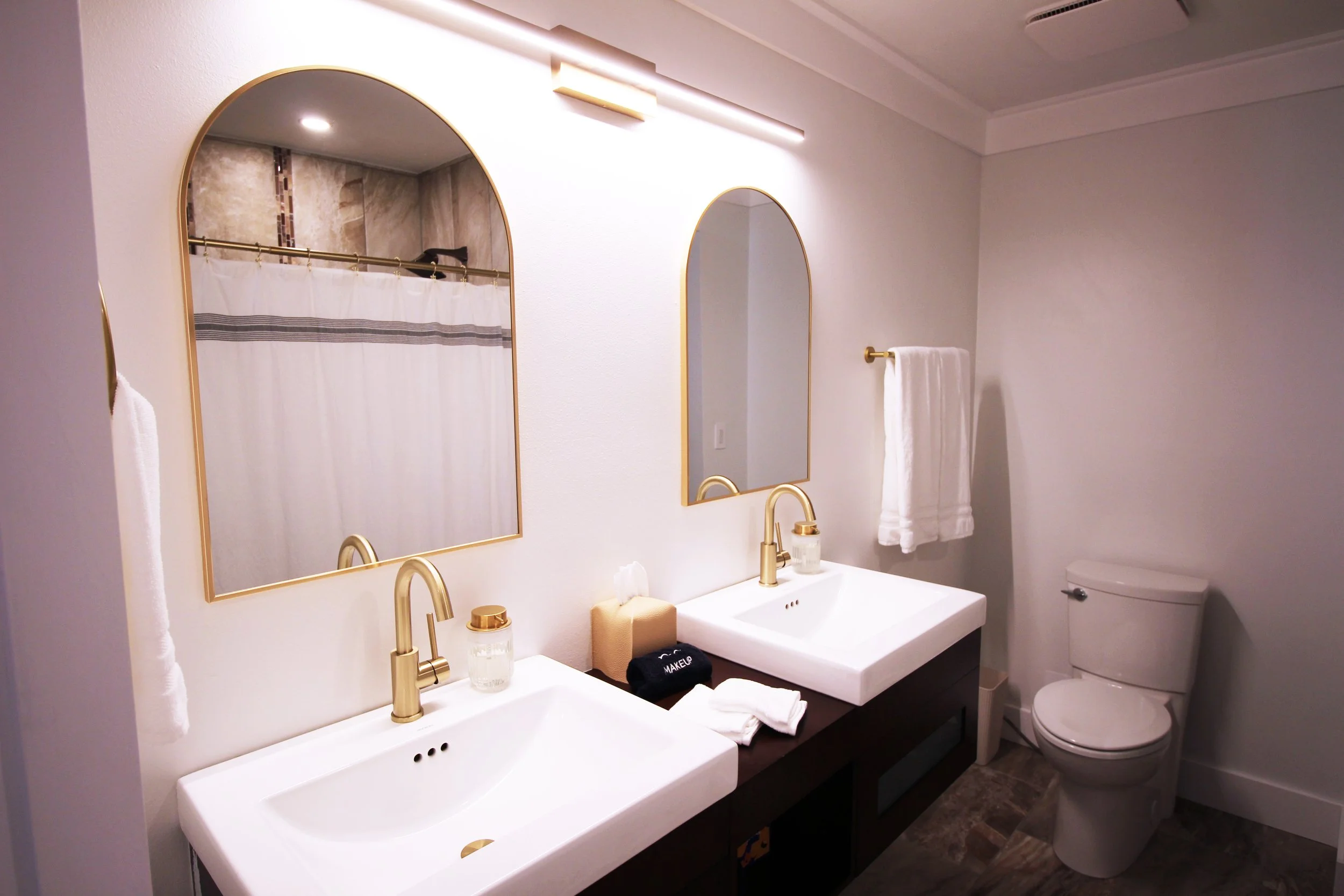 A large bathroom with white walls, double vanity sinks with matte gold fixtures, bright lighting, two arched mirrors, white towels, and a toilet.