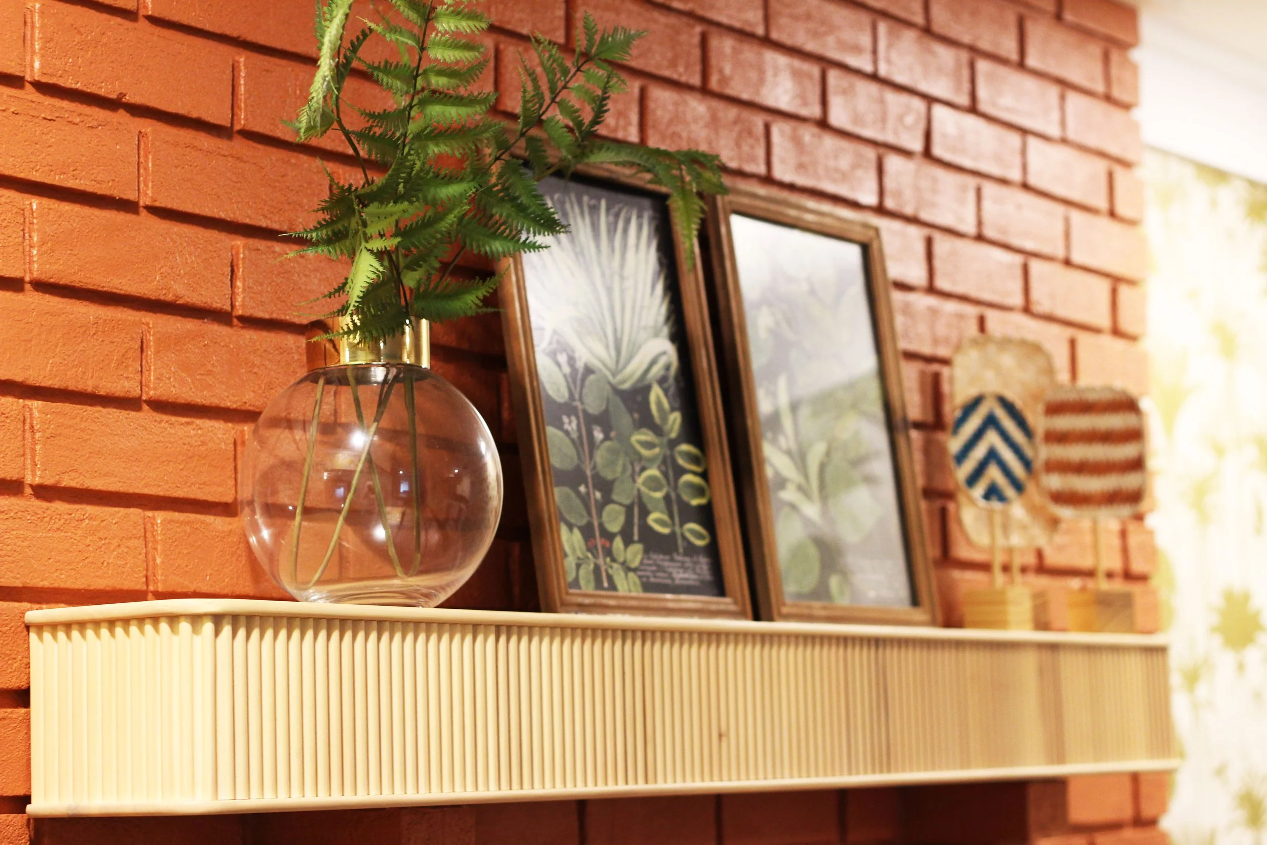 A decorative shelf with a glass vase of green fern leaves and framed botanical prints, mounted on a red brick wall with two small woven wall hangings in the background.