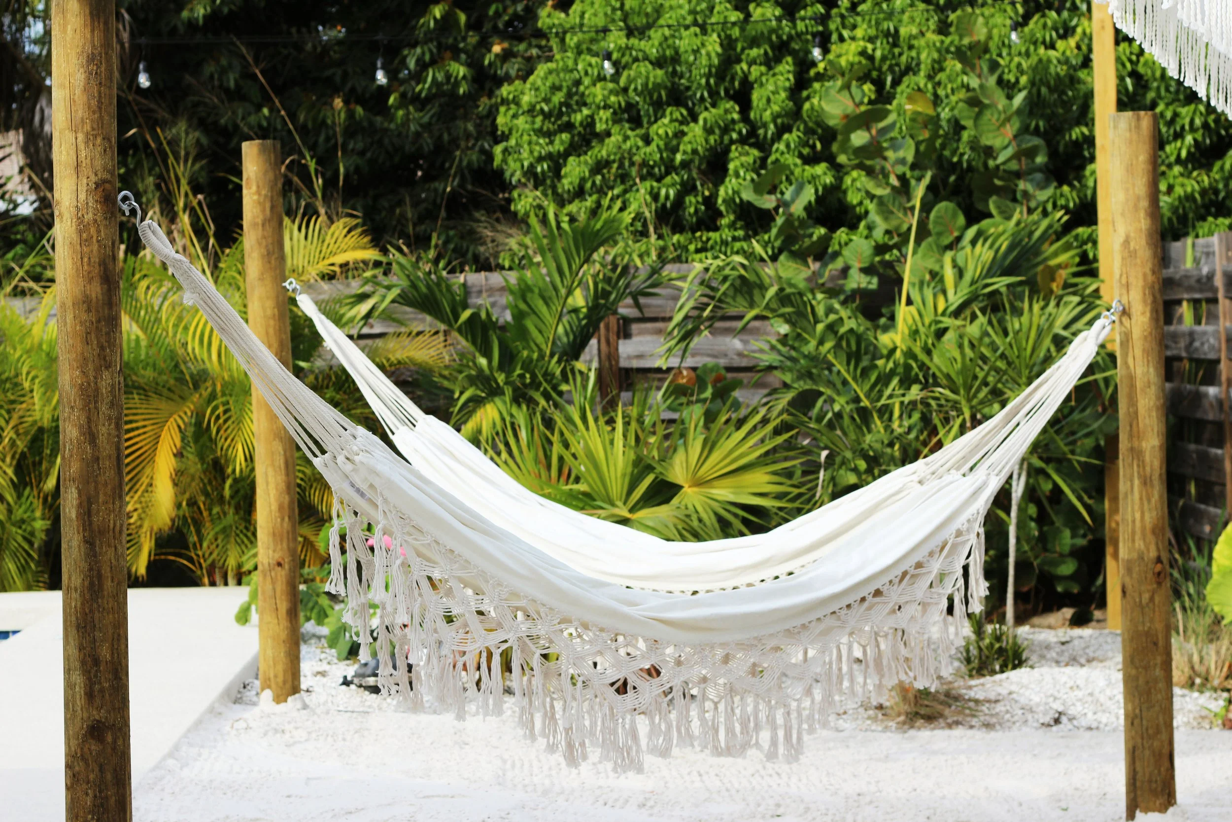 Two large, white hammock with boho style fringe details hanging between wooden posts over the white sand beach area, with lush green tropical plants in the background.