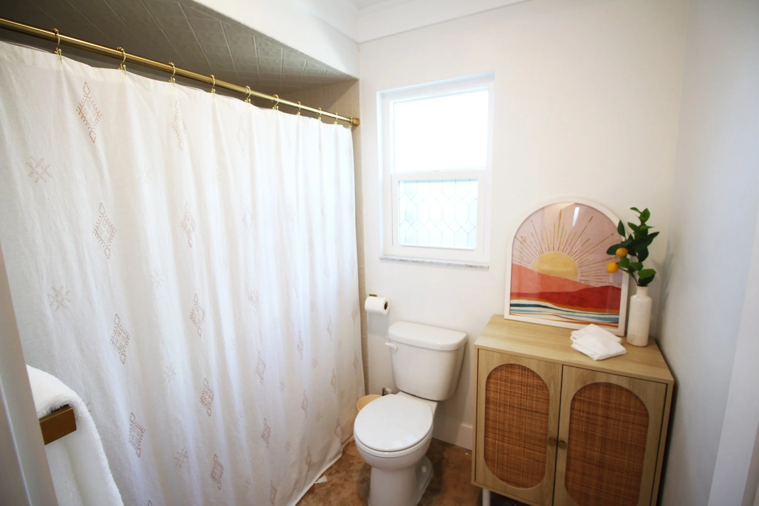 A bathroom with white walls, a large shower, a toilet, a small cabinet adorned with a decorative sunset picture and vase, and a small window for natural light.