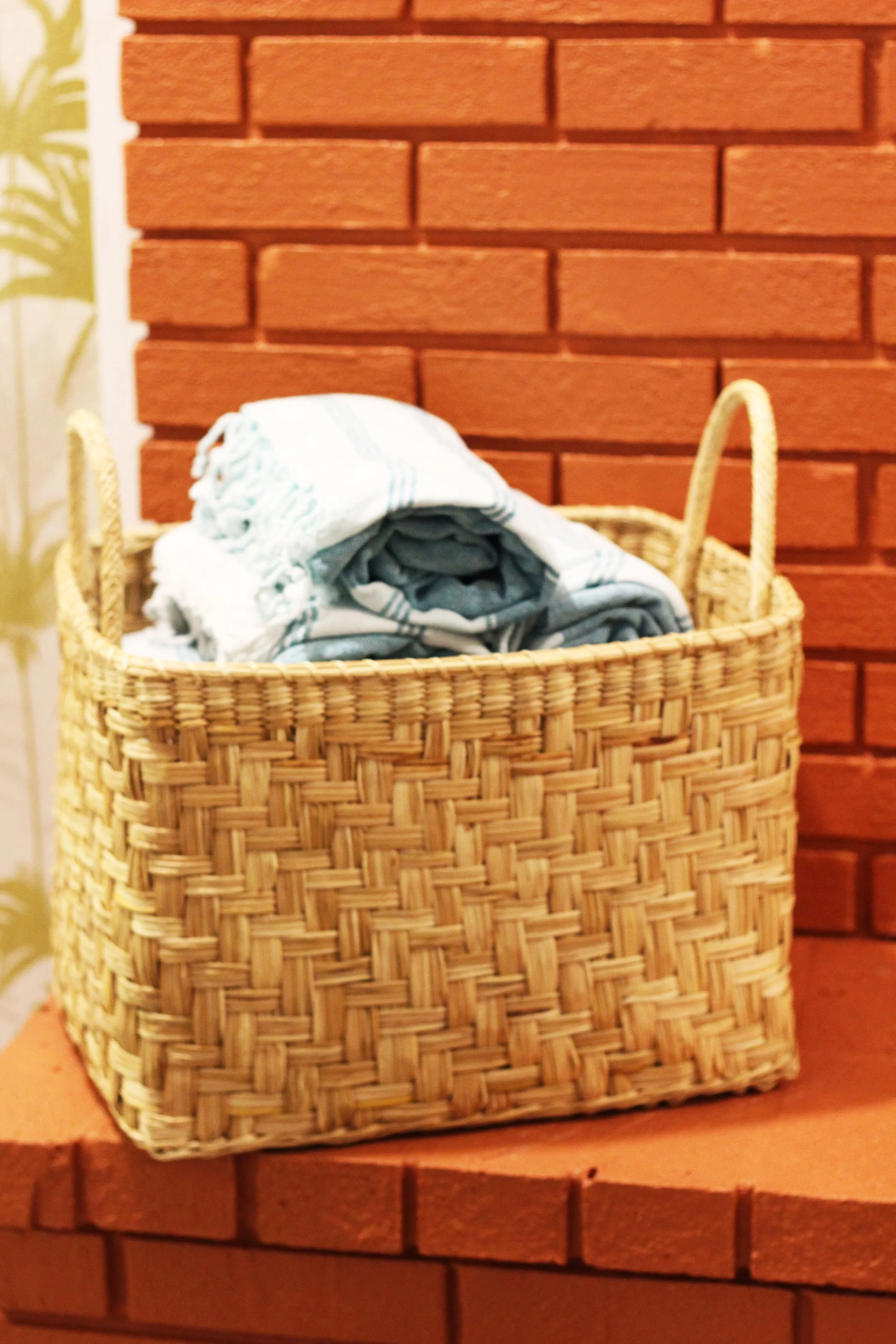 A close up view of a woven wicker basket with handles, filled with folded beach towels, sits on a brick fireplace mantel.