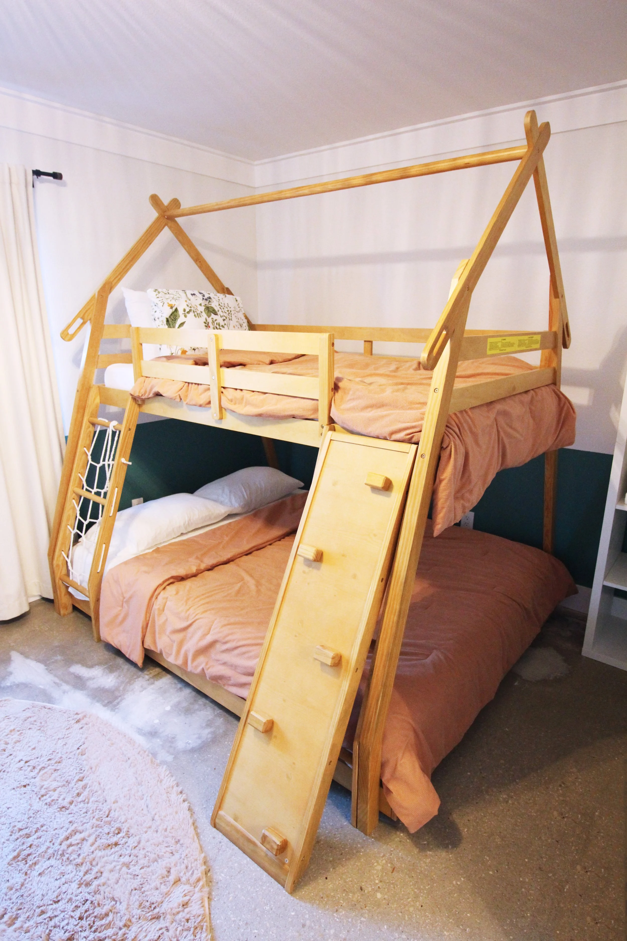 A bunk bed with a fun, house-shaped wooden frame, with a lower Queen bed and an upper Twin bed. The frame includes a ladder, rope wall, and climbing wall to make it fun for kids. The top bed has a guardrail.