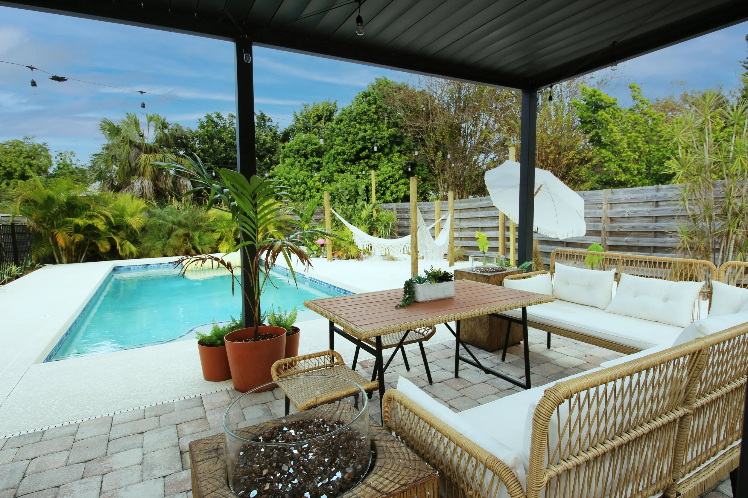 The backyard patio area with boho style outdoor furniture, a swimming pool, potted plants, tropical landscaping, a large white sand beach area, two hammocks, cafe lights, background of trees and blue sky.
