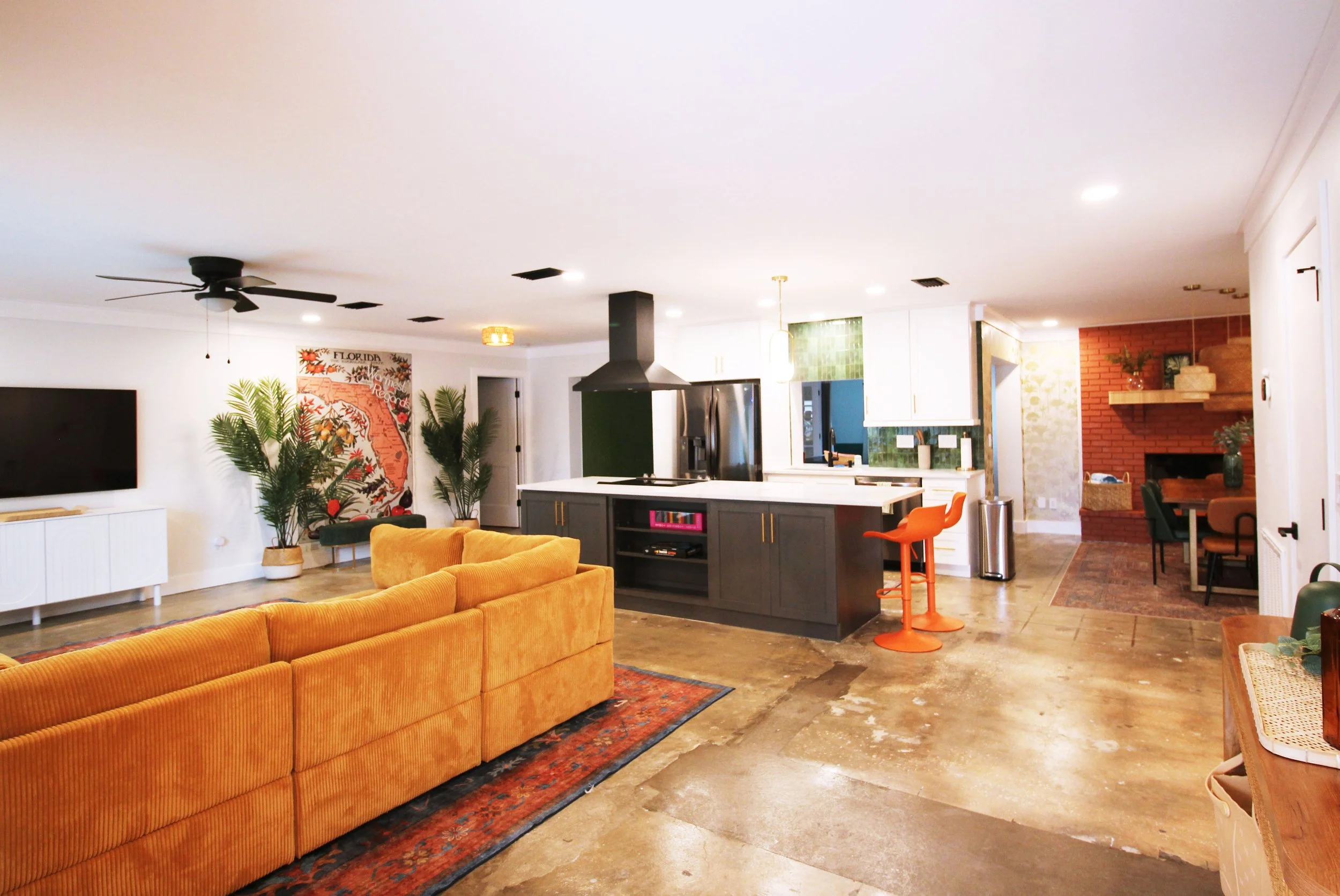 Open-concept living room and kitchen with retro yellow corduroy sofa, fun orange bar stools, tropical plants, and brick fireplace in the background.