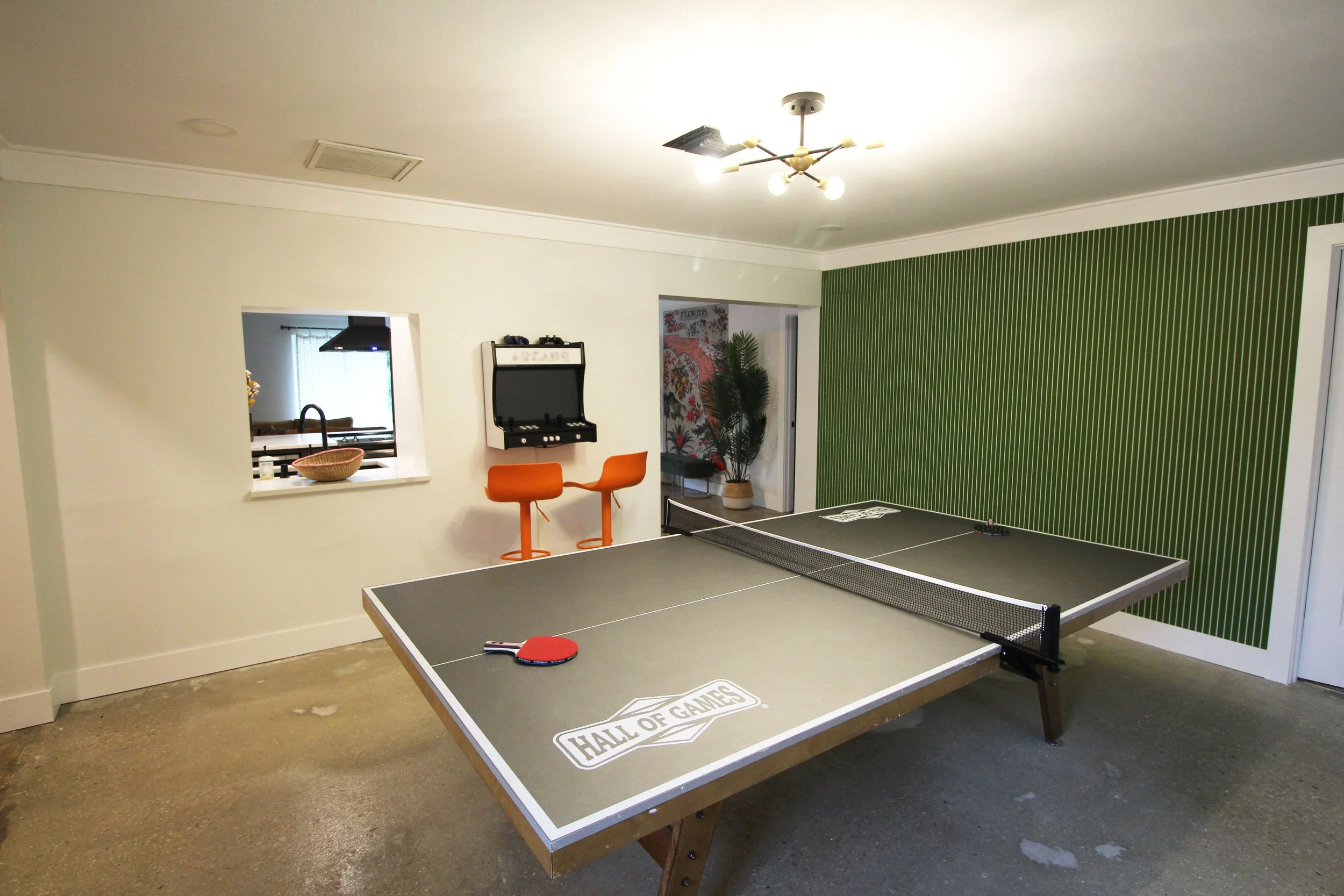 Indoor game room with large, wooden ping pong table, two fun orange bar stools, a wall mounted arcade machine with 6000+ games, and a pass through window to the kitchen.