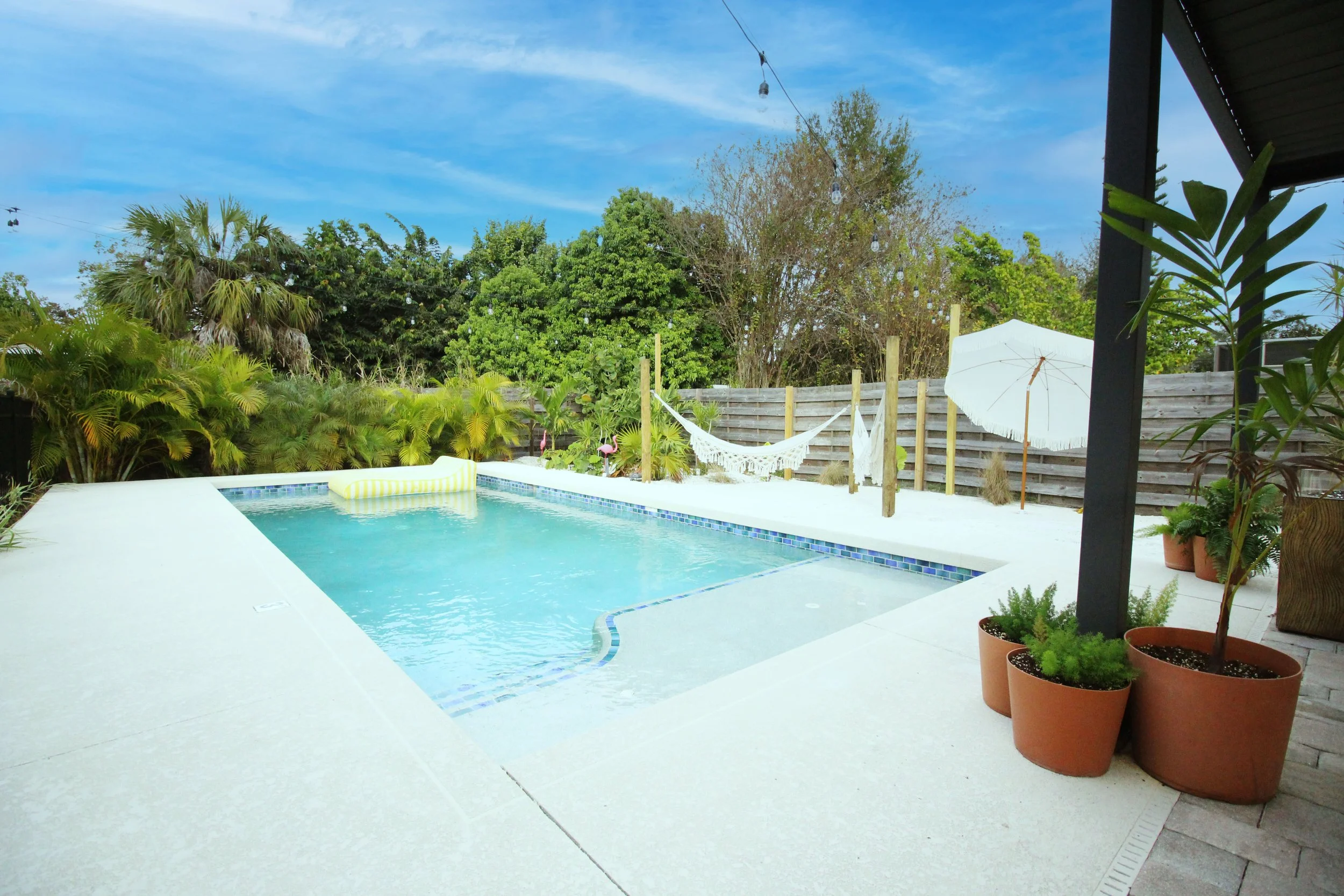A centered view of the backyard swimming pool with lounge float, white umbrella, potted plants, hammocks, fence, and trees under a blue sky with clouds in the background.