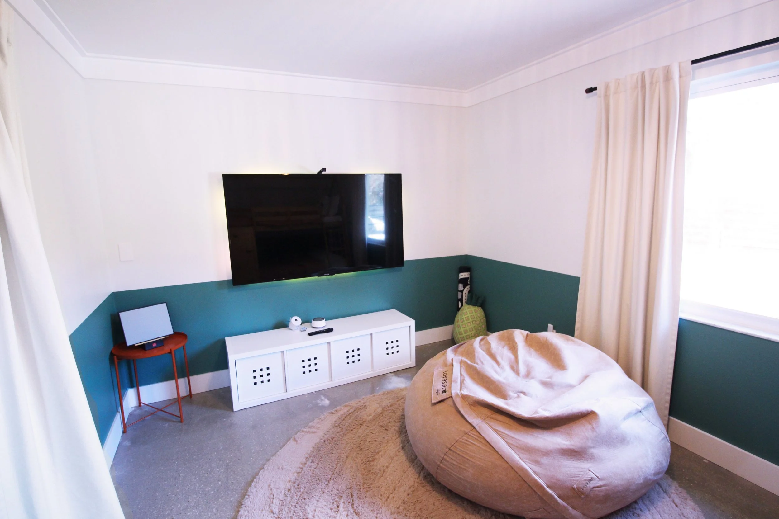 A bedroom with two-tone white and green walls, a wall mounted smart TV , a white TV console, a plush round beige bean bag chair, a small orange side table and curtains covering a window with natural light.
