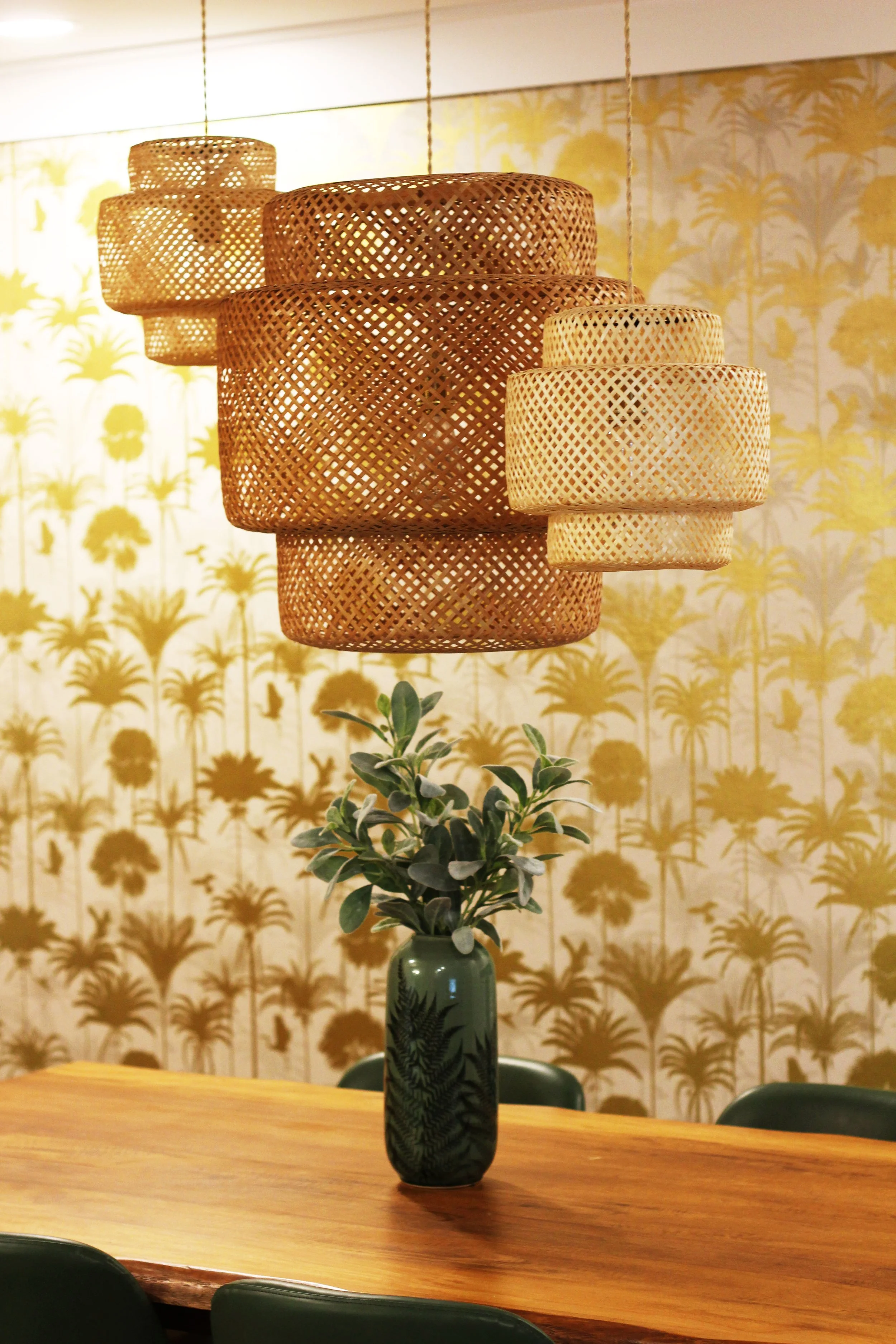 A close up view of a green vase with leafy plant on the dining table, decorative woven hanging lamps above, patterned tropical-themed wallpaper with palm trees in the background.