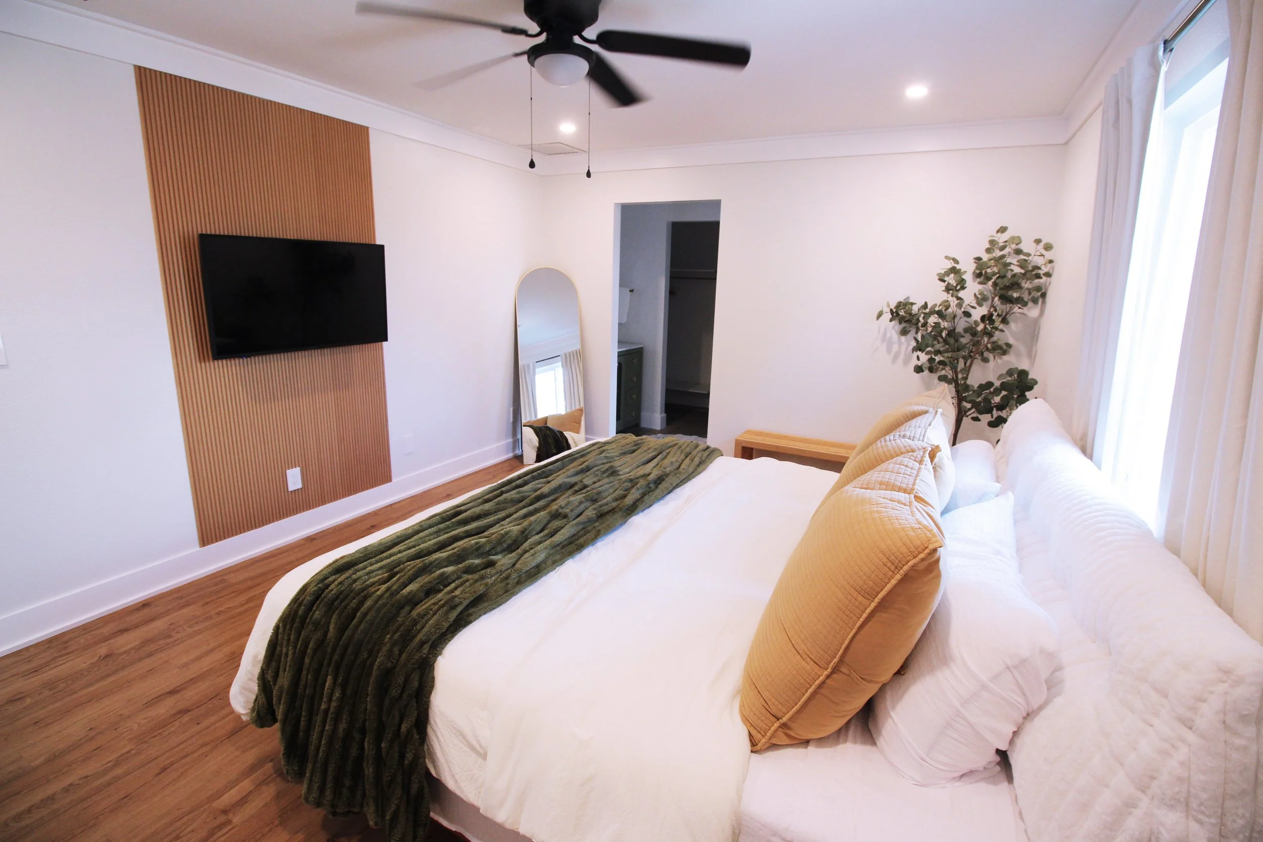 A large bedroom with white walls, hardwood floors, a King bed , hardwood floors, a wall-mounted smart TV on a light wood tambour accent wall, a tall mirror leaning against the wall, a window with curtains, and a ceiling fan.