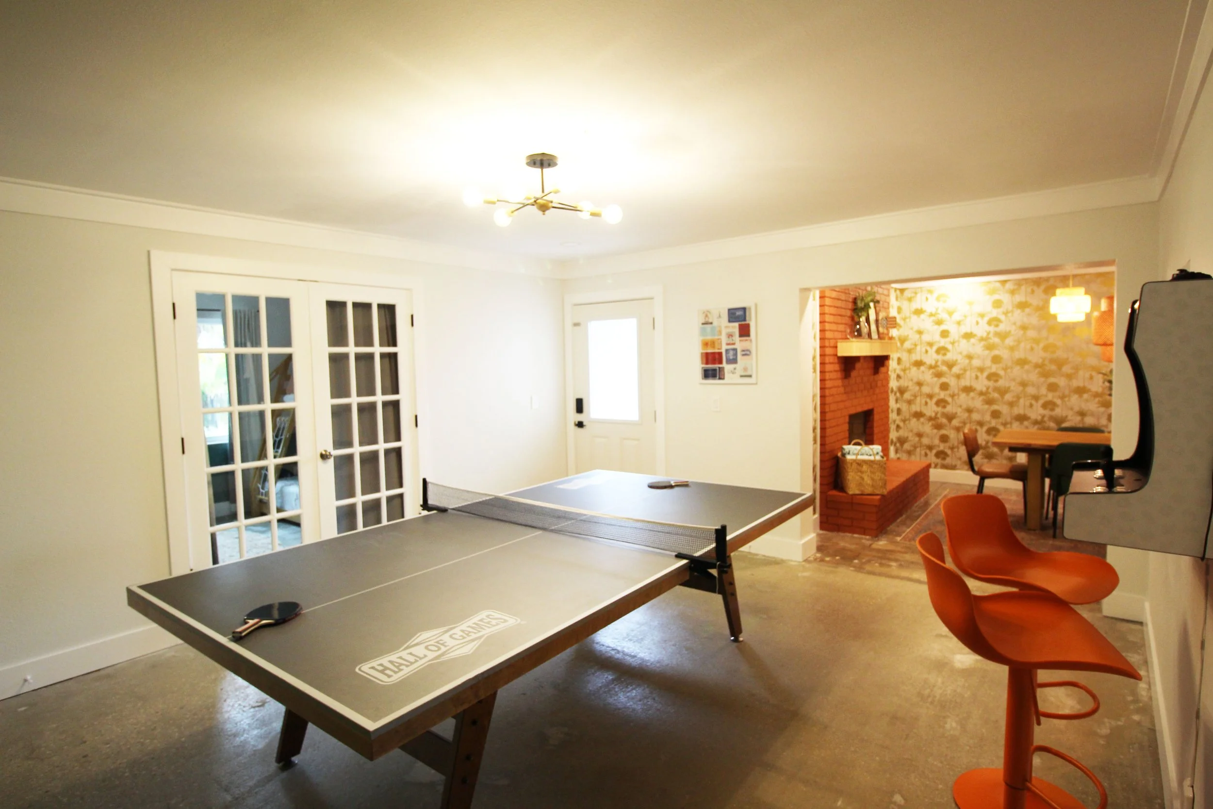 Another view of the indoor game room with large, wooden ping pong table, two fun orange bar stools, a wall mounted arcade machine with 6000+ games, and an entrance into the dining room.