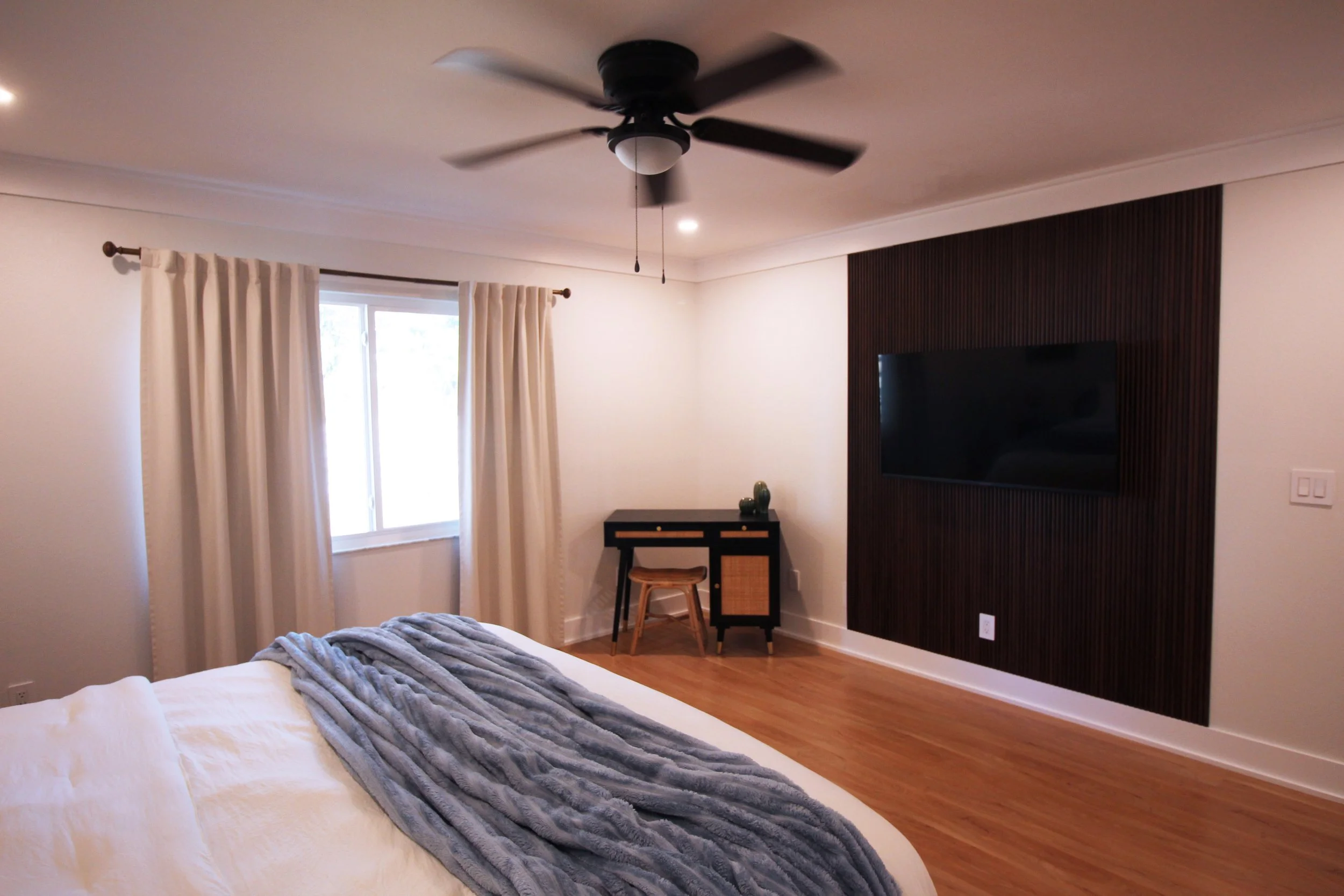 A large bedroom with white walls, hardwood floors, a King bed with a white sheets and a blue throw blanket, window with cream curtains, a black desk with a small stool, a dark wood tambour accent wall with a mounted smart TV and ceiling fan.
