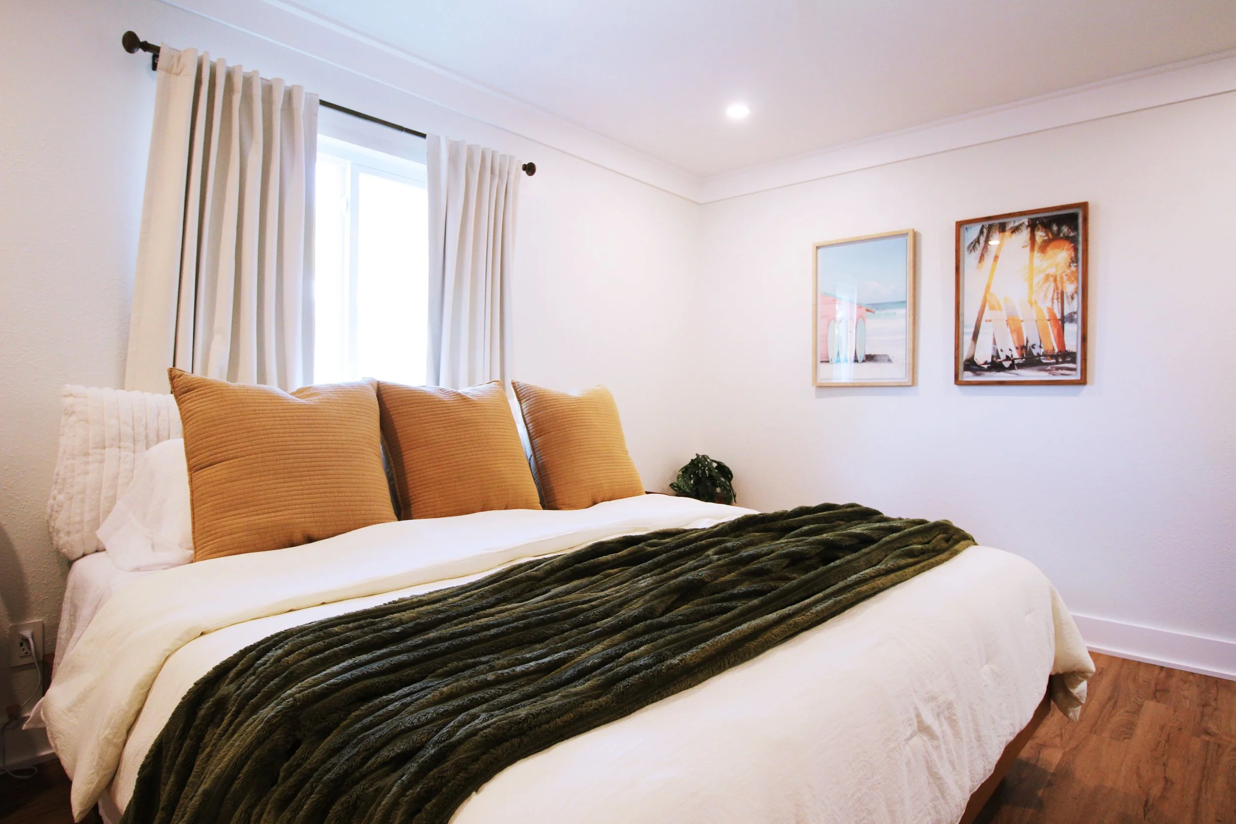 A large bedroom with white walls, hardwood floors, a King bed, beige pillows, white sheets, a dark green throw blanket. A window with white curtains, two framed seaside paintings on the wall, and a small plant in the corner.