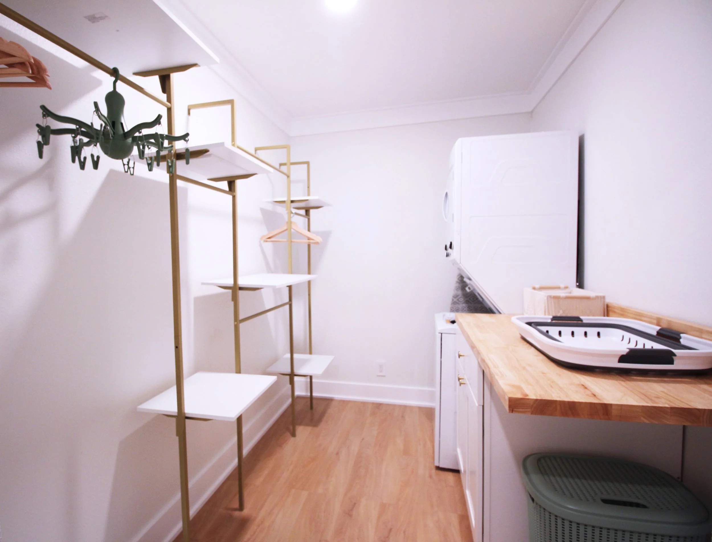A large, walk-in closet with stacked washer and dryer, white cabinet with butcherblock countertop, and modern white shelving with hangers. 