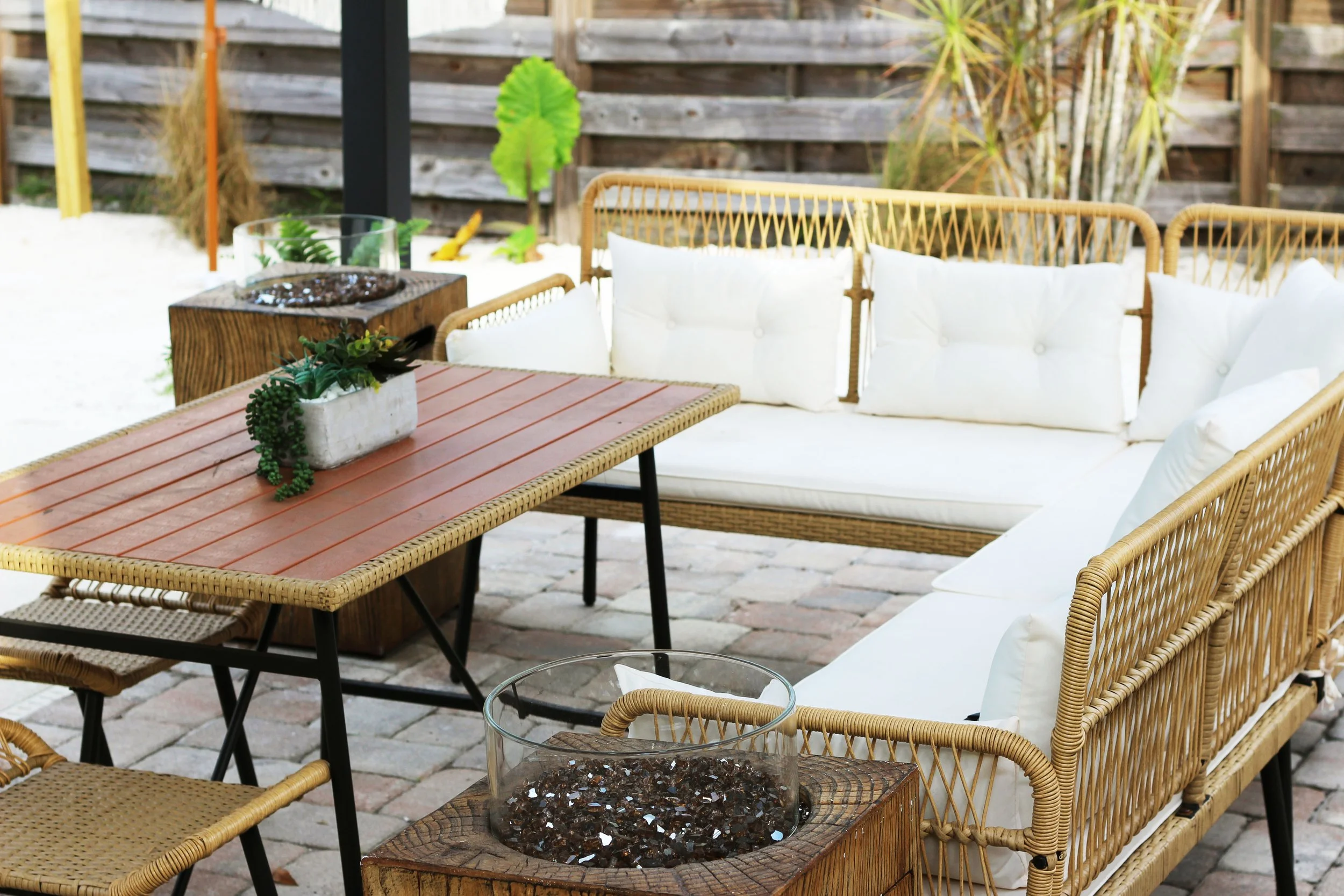 Outdoor patio furniture set with a boho style wicker sofa, dining table with a plant centerpiece, and side table firepits.