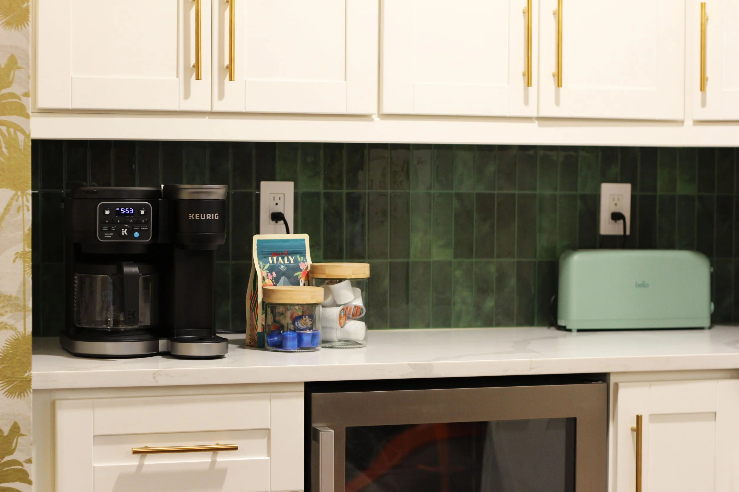 A butler's pantry area, with white quartz countertop, a black Keurig coffee maker, a box of accompaniments, two jars with wooden lids, a teal toaster, and two electrical outlets on a green tiled backsplash.