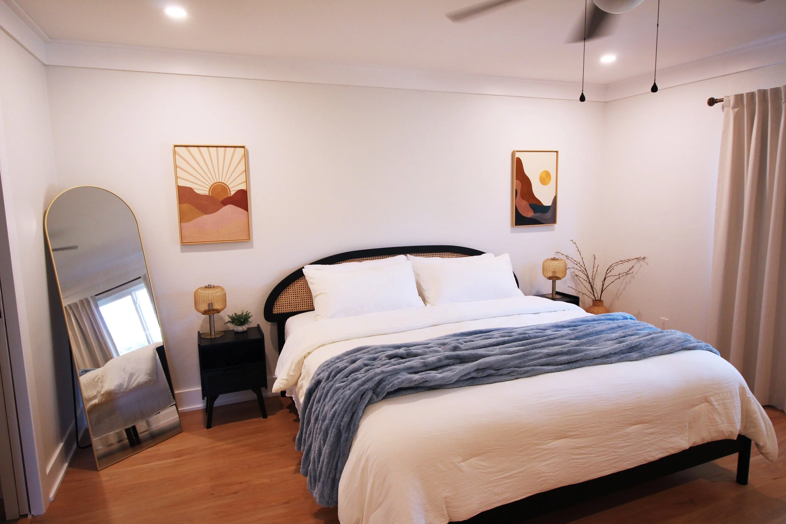 A large bedroom with white walls, hardwood floors, a King bed with white linens, pillows, a blue throw blanket, and a black headboard with boho caning accents. There are two black nightstands with lamps and a tall, curved standing mirror