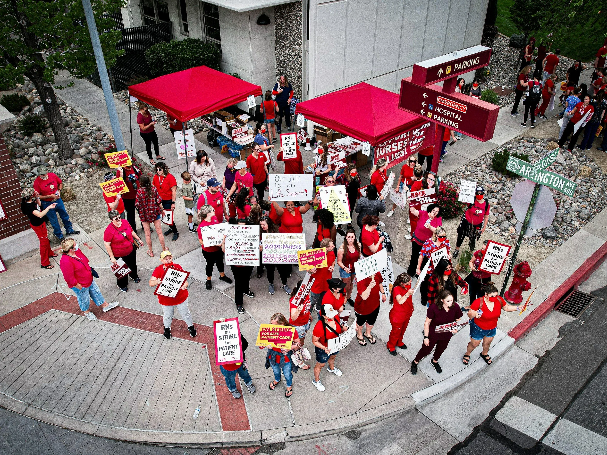 A crowd of people participating in a protest or strike outside a building. Many are holding signs with messages supporting nurses and patient care. The group mostly wears red and some signs reference nurses on strike and safe patient care.