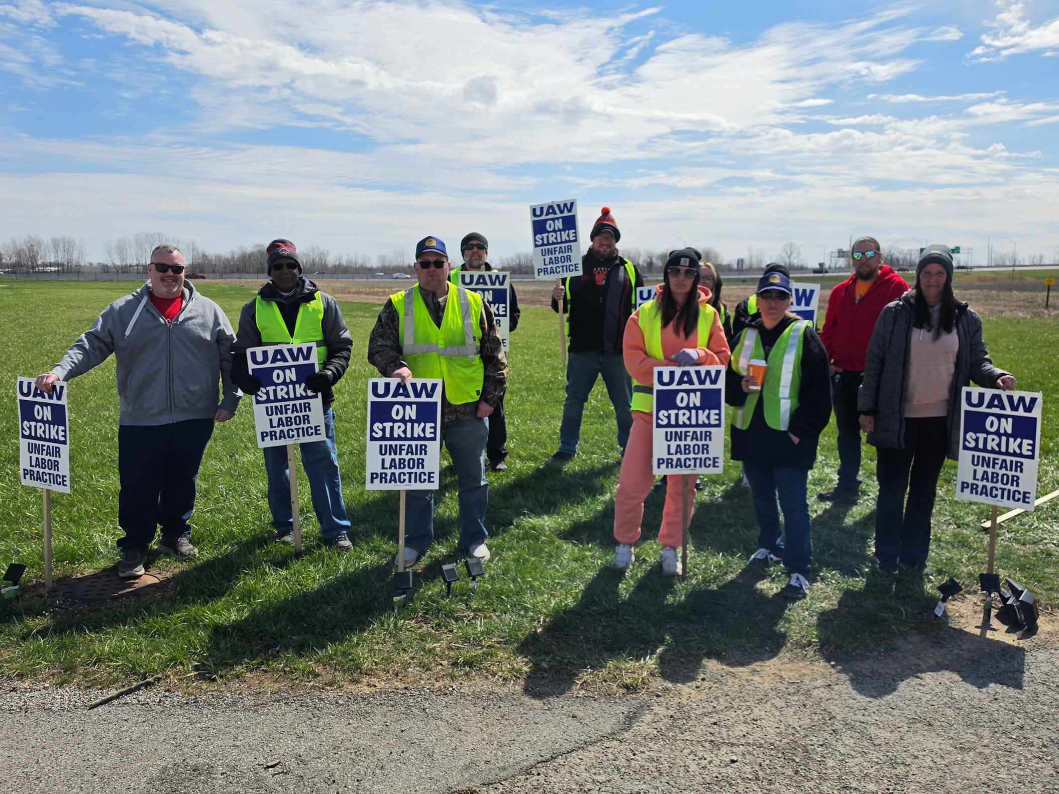 A group of people protesting for workers' rights, holding signs that read 'UAW ON STRIKE' and 'UNFAIR LABOR PRACTICE.' They are standing outdoors in a grassy area on a partly cloudy day.