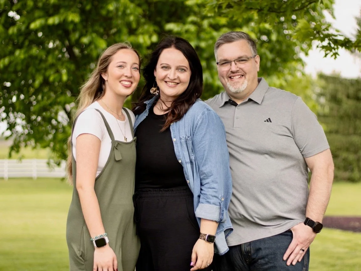 Three happy people standing outdoors in a park with green trees in the background.