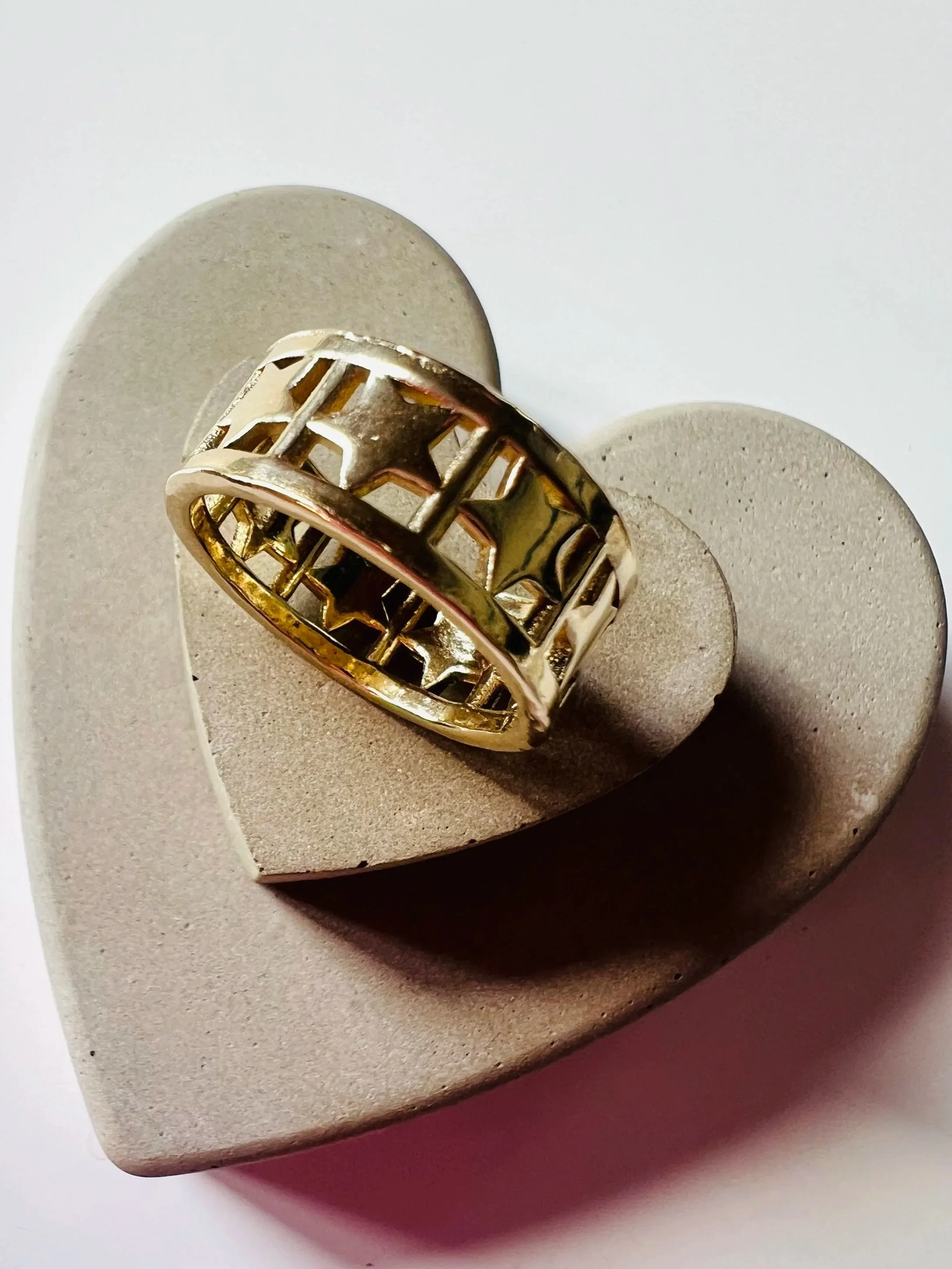 A gold ring with star and shaped cutouts, resting on a heart-shaped concrete or plaster surface.