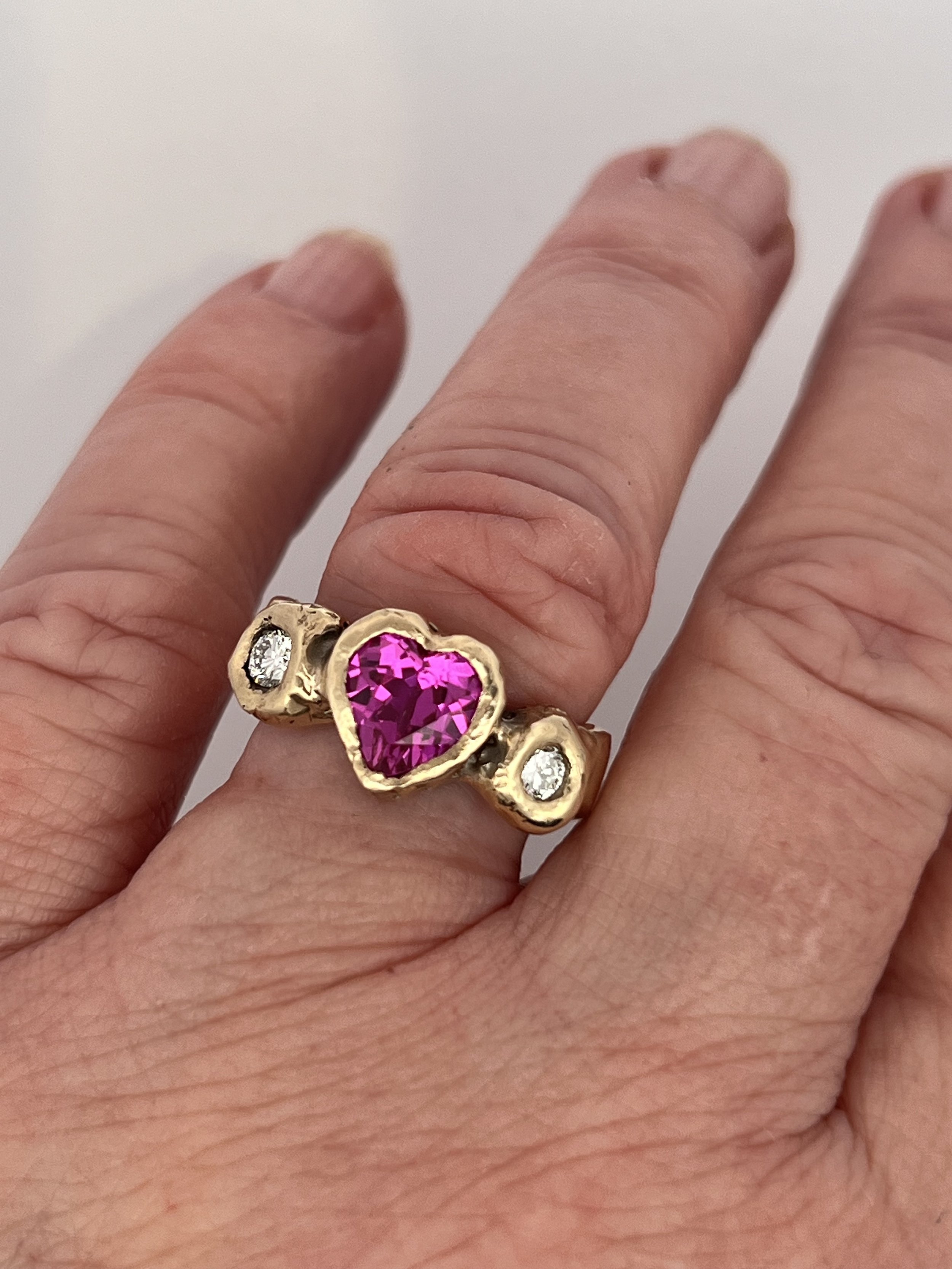 Gold ring with heart-shaped opening, placed on a heart-shaped gray surface. Two small clear gemstones and a small red heart-shaped gemstone are also on the surface.