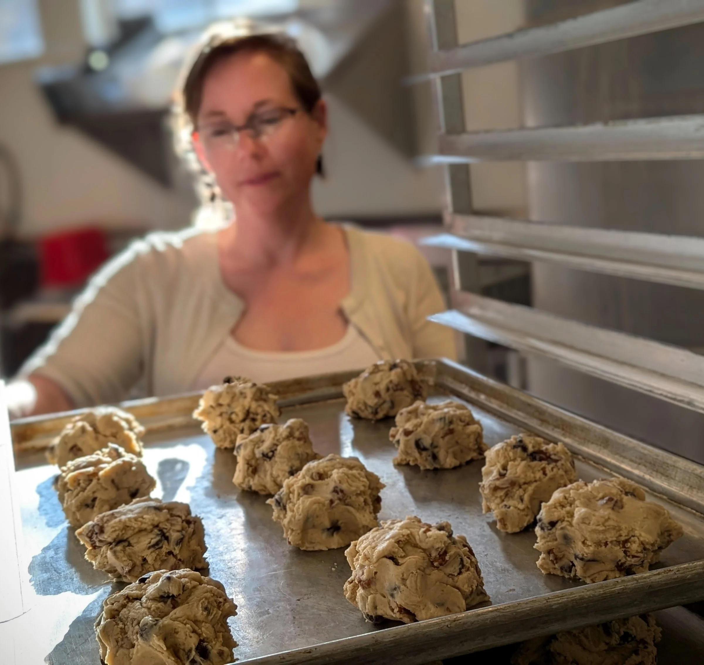 A woman with short brown hair and glasses stands behind a baking sheet with cookie dough, baking chocolate chip cookies fresh from the oven.