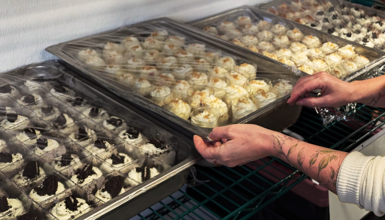 Person opening a clear plastic lid on large trays of baked desserts, which include cookies and cupcake-like treats with white frosting and toppings.
