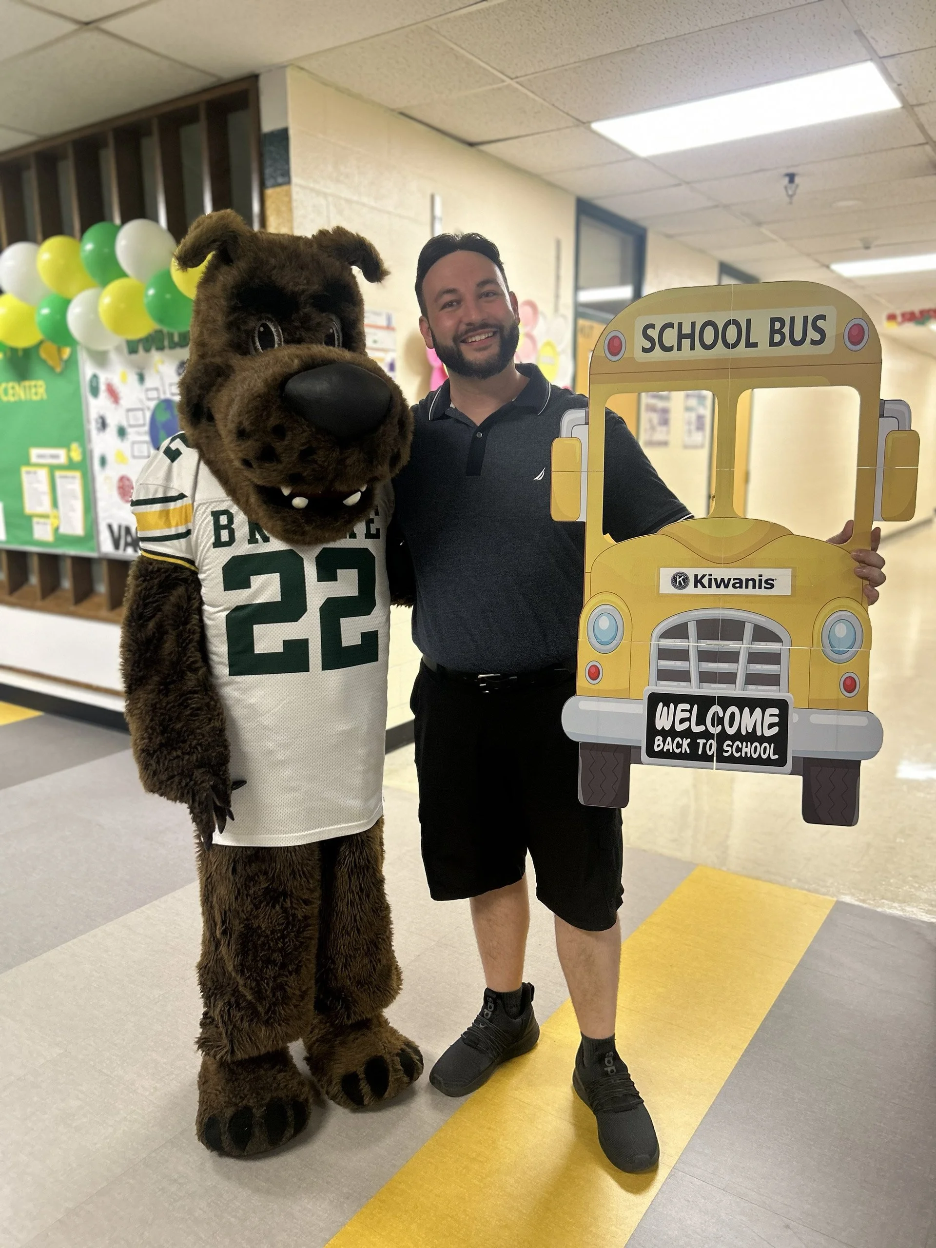 A man dressed in casual clothing standing next to a mascot in a bear costume wearing a football jersey, both smiling, in a school hallway decorated with balloons and a bulletin board. The man is holding a large cardboard cutout of a school bus with a