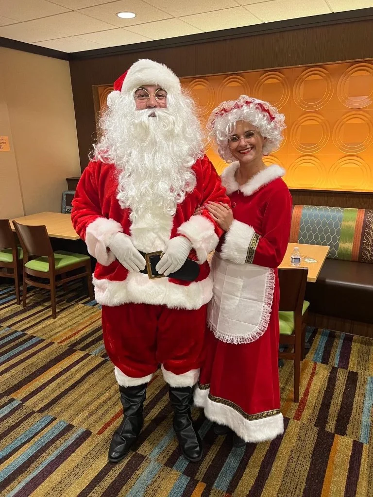 People dressed as Santa Claus and Mrs. Claus standing together in a restaurant or cafe, smiling, with a table and chairs visible in the background.