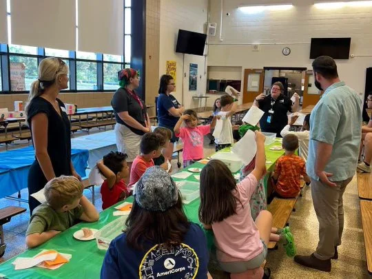 Children and adults gathered around a long table in a classroom or community center, participating in a group activity with papers and supplies, with large windows, TVs, and a teacher or facilitator at the front.