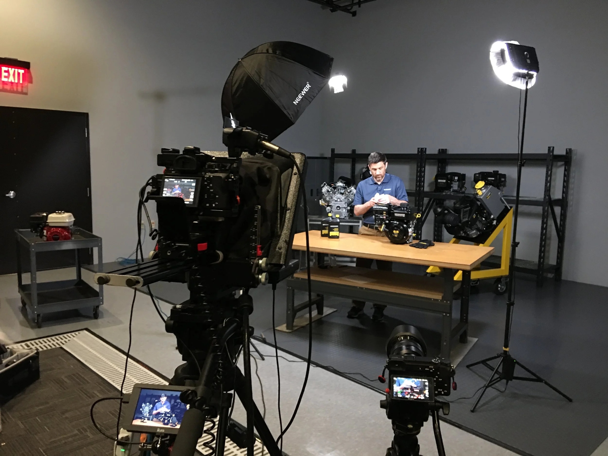 A man with a beard in a blue shirt is standing at a table with various camera equipment, including a camera, a large camera lens mounted on a stand, and several lights and reflectors. The room has grey walls, shelves in the background, and a black door on the left side. Camera and lighting equipment are set up for filming or photography.
