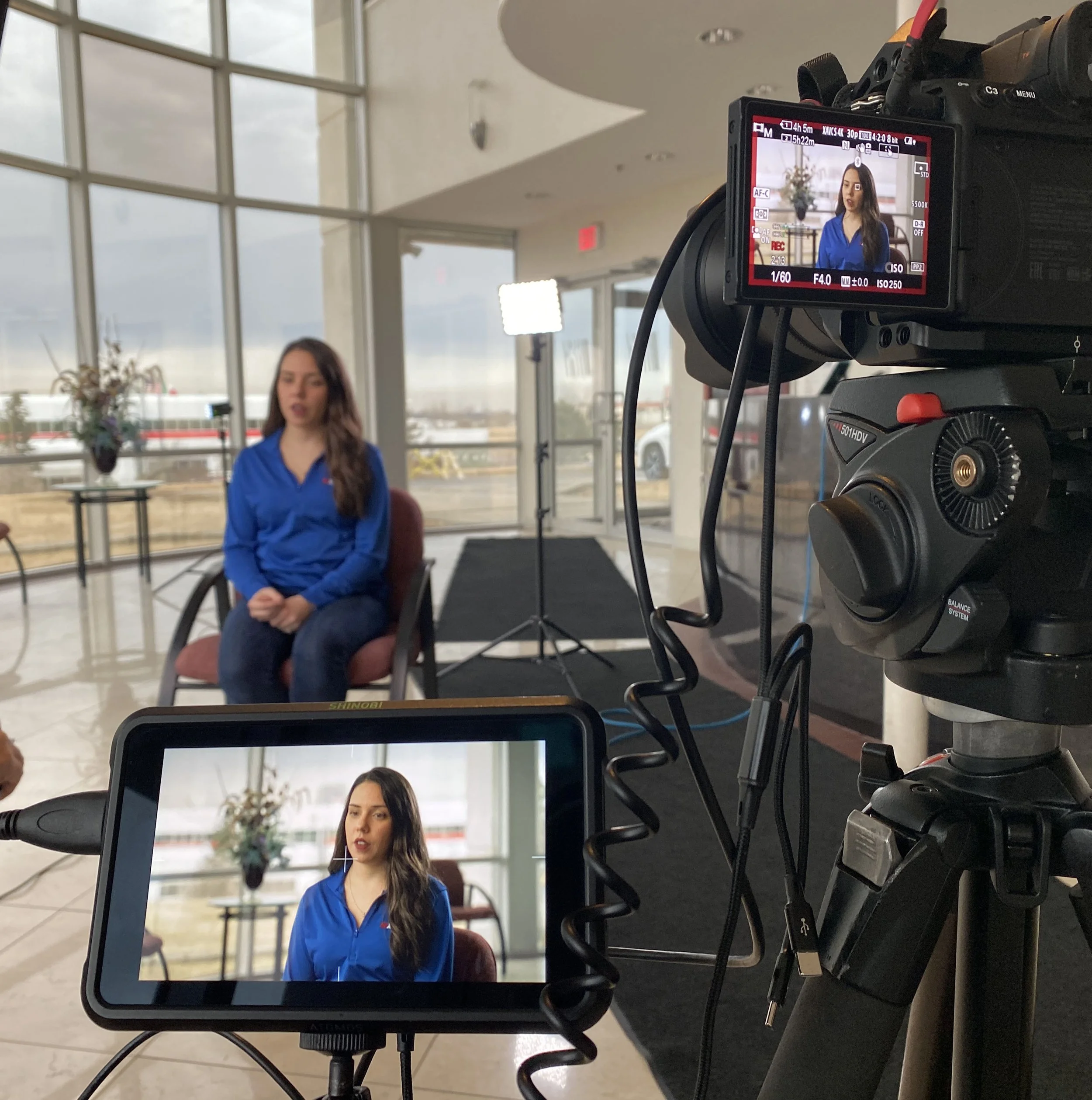 A woman with long brown hair wearing a blue blouse is sitting in front of a camera in an indoor setting, with large windows in the background showing the sky and some buildings.
