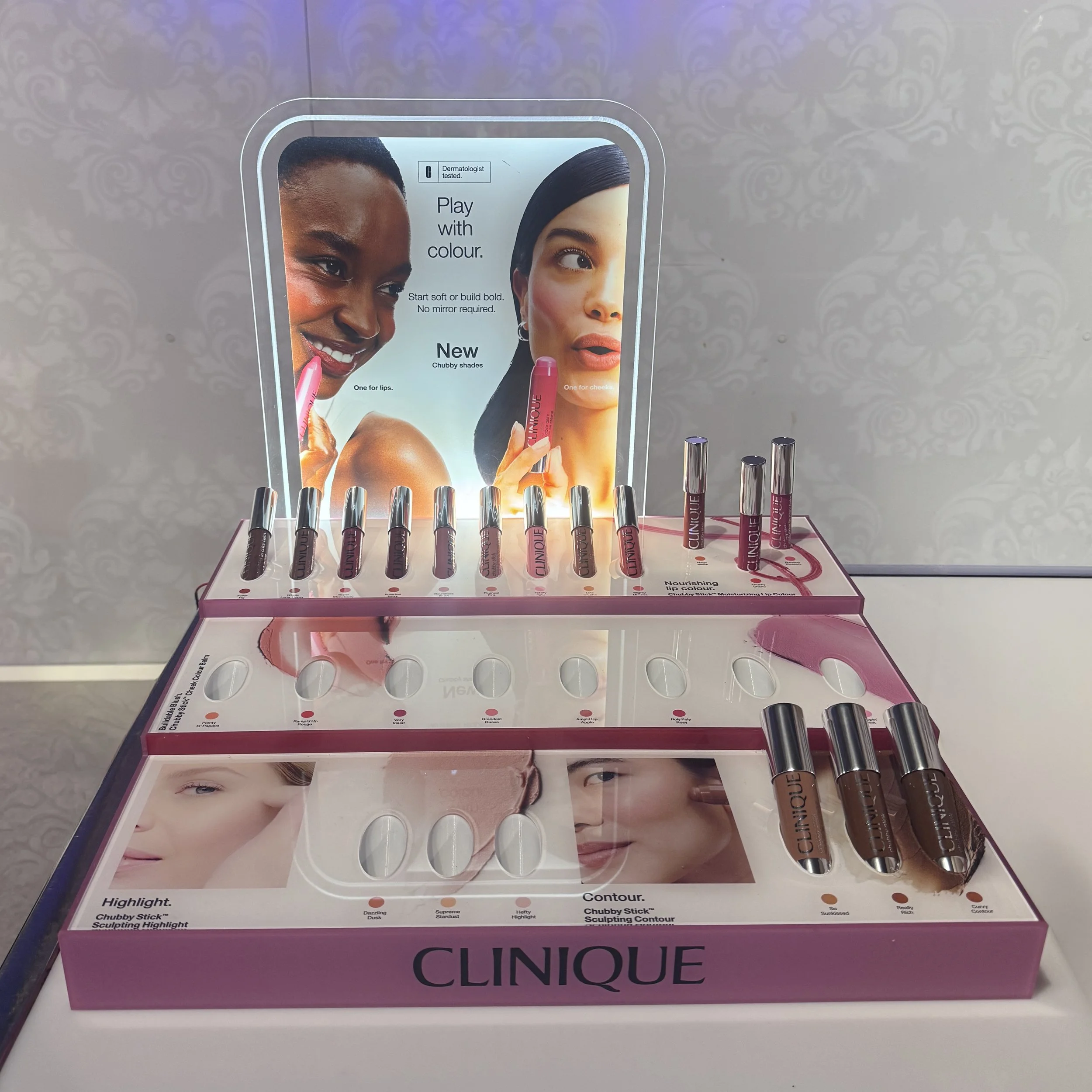 Display of Clinique makeup products with promotional poster in the background showing two women with colorful makeup.