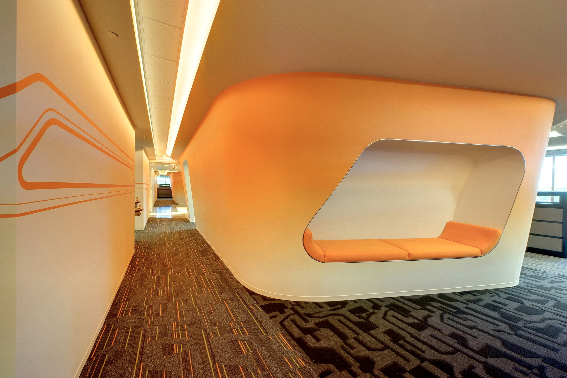 Modern indoor corridor with curved orange and white walls, featuring a built-in seating nook with orange cushions, and patterned carpet flooring.