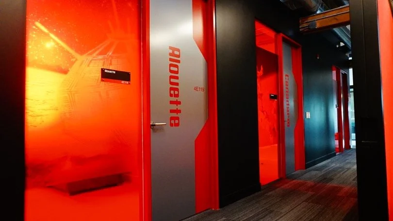 Hallway of an office or gaming space with doors labeled 'AIOUI:18' in red and gray, featuring a futuristic, tech-inspired design with a vibrant red and black color scheme.