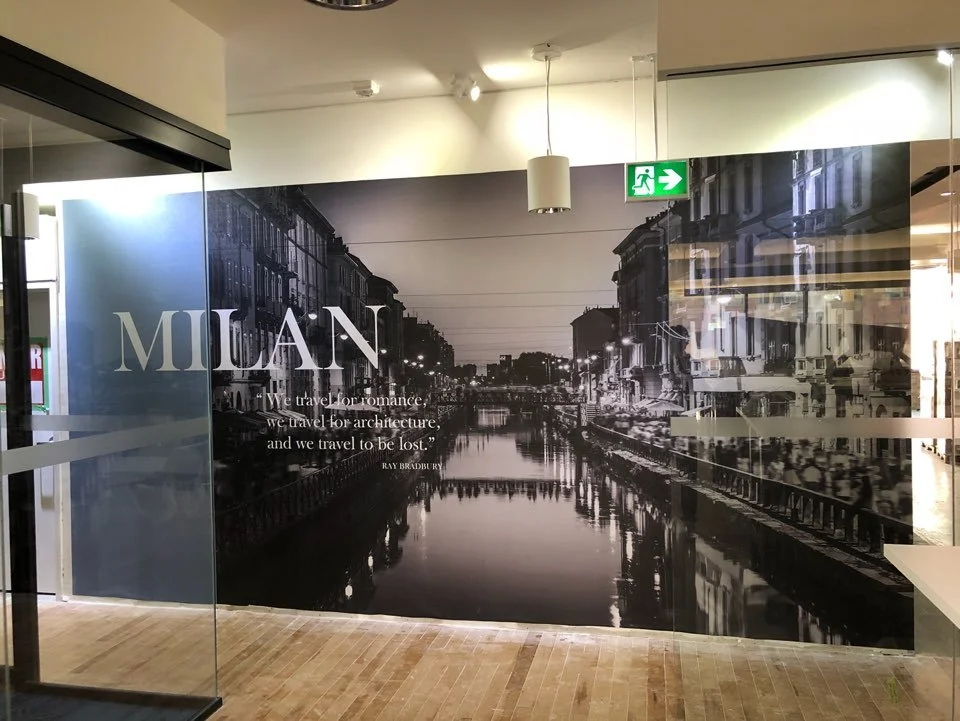 ~Black and white mural of a city canal with buildings on both sides, and lights reflecting on the water. There is a quote from Ray Bradbury: "We travel for romance, we travel for architecture, and we travel to be lost."
