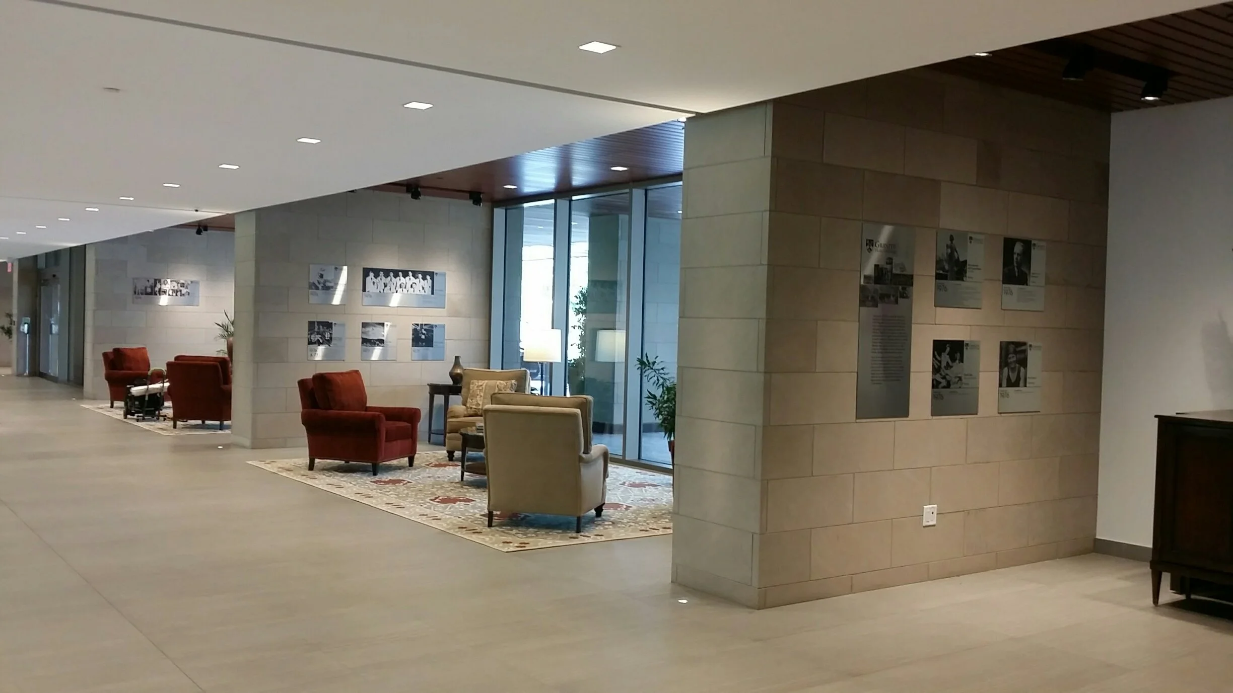 ~A spacious, well-lit lobby with seating areas featuring red and beige armchairs, a wooden table, lamps, potted plants, and wall displays of photographs and information.