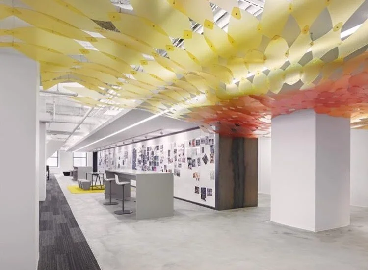 ~Modern office space with colorful hanging ceiling art, white walls covered with photos and artworks, a white counter with barstools, and a seating area with chairs and a yellow rug.