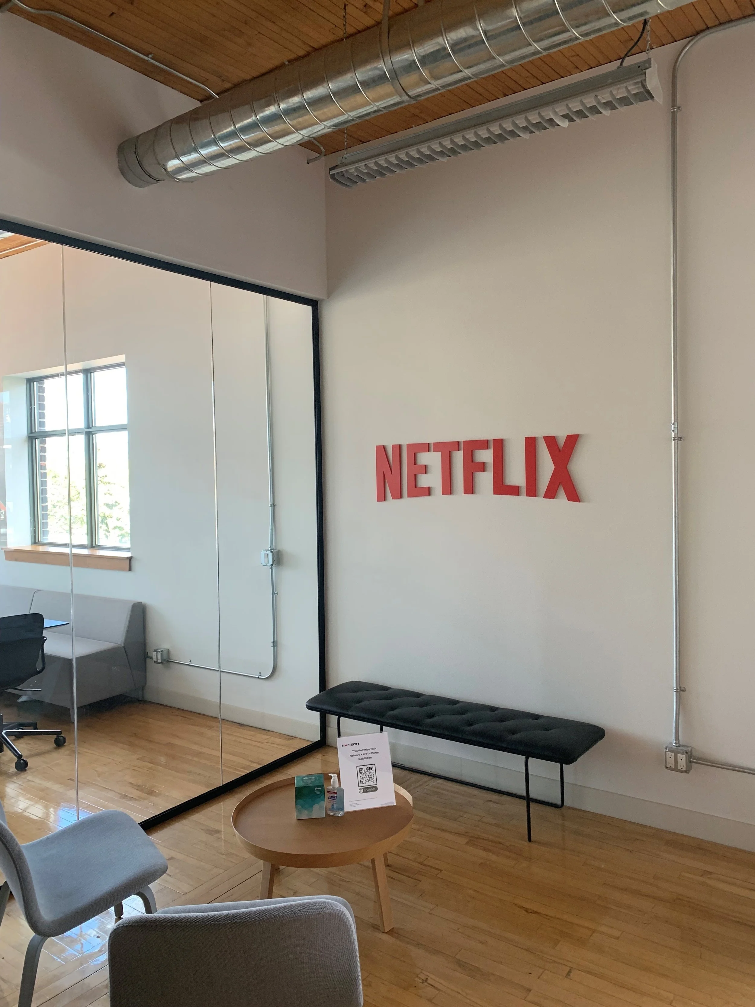 ~An interior office space with a Netflix logo on the wall, a black bench, a small round wooden table with hand sanitizer and a QR code sign, and a glass wall reflecting a window and office furniture.