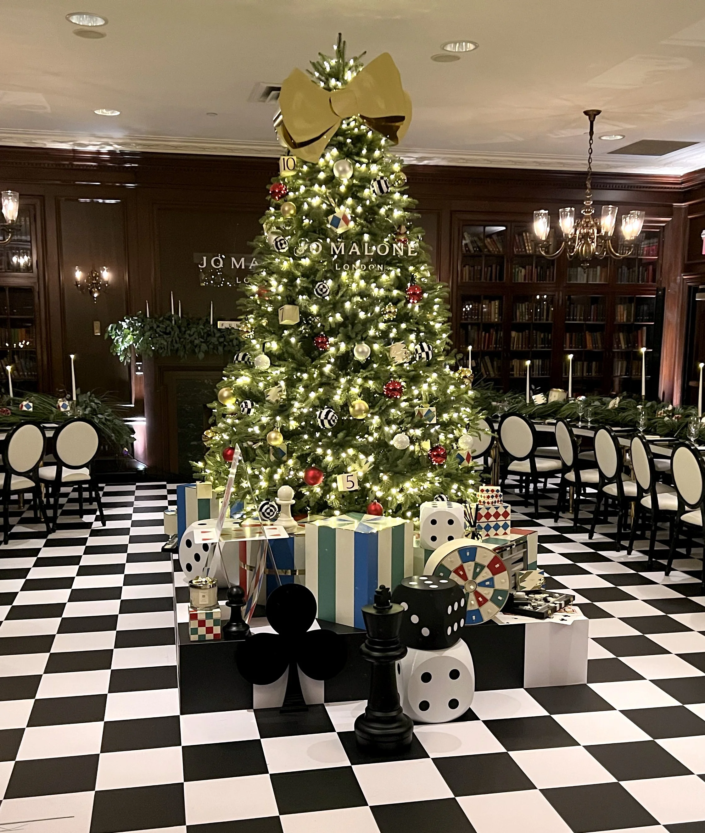 Decorated Christmas tree with ornaments, presents, and game pieces in a festive room with black-and-white checkered floor and wood-paneled walls.
