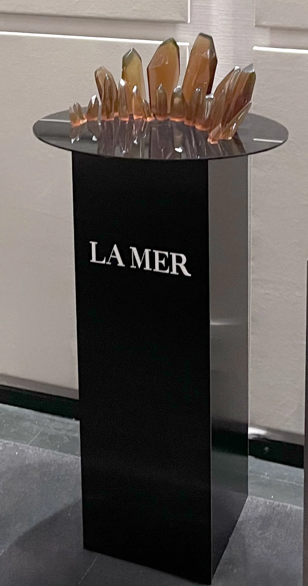 A black display stand with the logo 'LA MER' in white letters, featuring a decorative sculpture made of multiple amber-colored crystals on top.