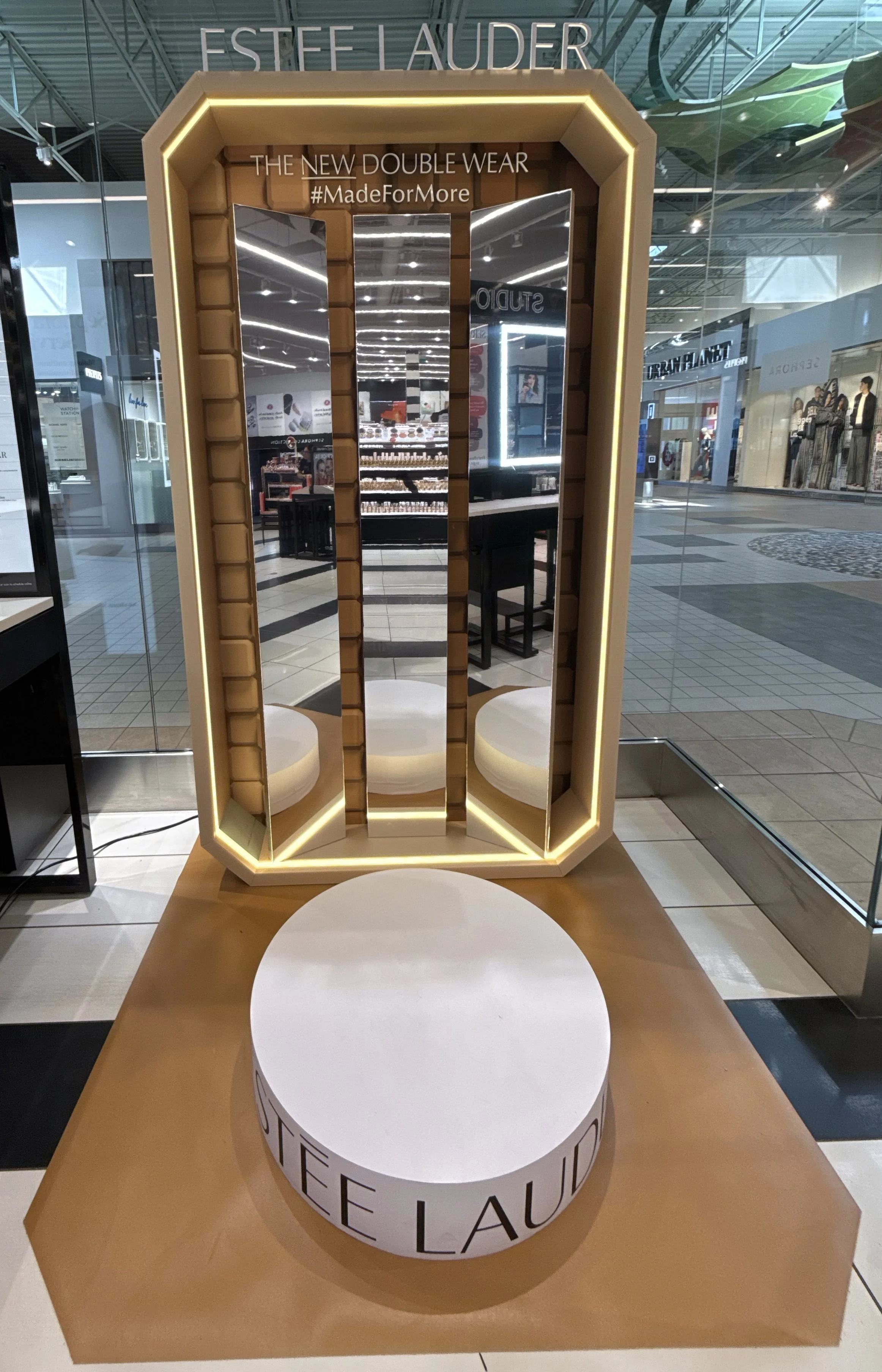 A modern Estée Lauder display with a large mirror and built-in lighting, showcasing the brand's new Double Wear makeup collection, inside a mall.